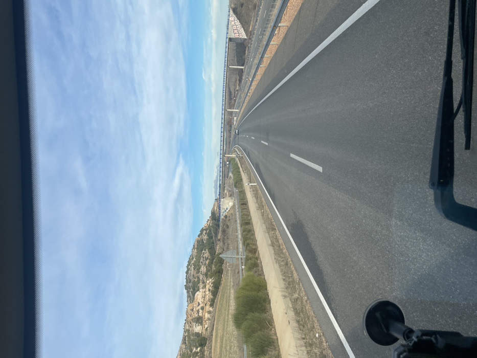 A straight, multi-lane highway stretches into the distance under a partly cloudy sky. A tall bridge is visible in the background, running parallel to the highway. Adjacent to the road is a hillside with sparse vegetation. The view is captured from inside a vehicle, with a windshield wiper and side mirror visible in the frame. The asphalt surface appears smooth and well-maintained, marked with white lane dividers.