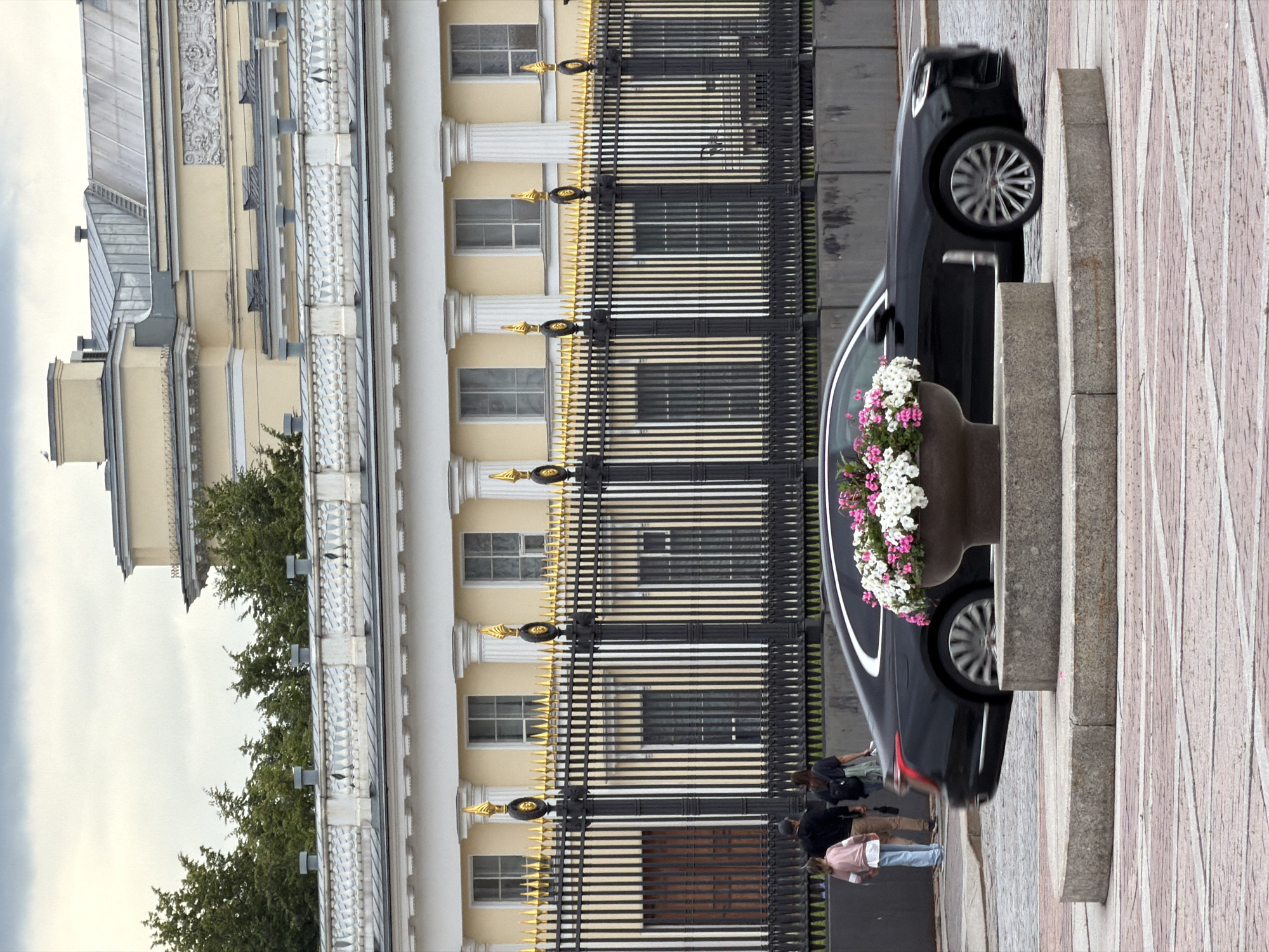 The image depicts a street scene in the city of Lednevo, Russia. A black luxury sedan is captured in motion, its wheels blurred, indicating movement. The car is positioned in the foreground, partially obscuring a large, ornate planter filled with pink and white flowers. Behind the car and planter, a black metal fence with gold decorative elements runs horizontally across the middle ground. Beyond the fence, a grand pale yellow building with white columns and multiple windows is visible. In the far background, trees and a cloudy sky complete the scene. To the left of the fence, three individuals are walking: a young girl in a pink top and blue jeans, a man in a dark shirt and brown pants, and a woman in a dark shirt and lighter colored pants carrying a bag. The ground in the foreground is paved with reddish-brown and grey interlocking stones.