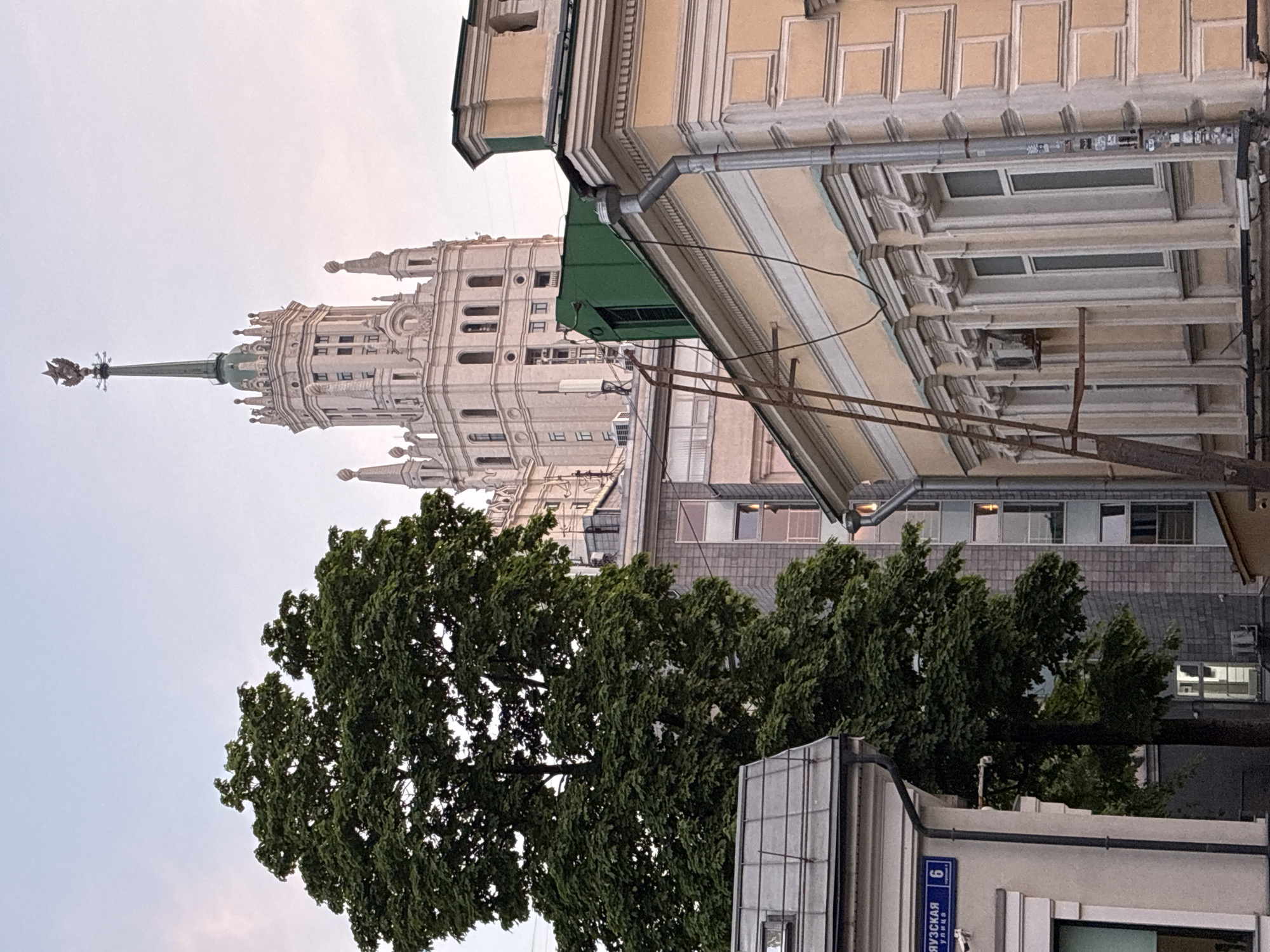 The image captures a view in Moscow, Russia, featuring a Stalinist skyscraper amidst trees and other buildings. The skyscraper, recognizable by its ornate design and spire topped with a Soviet star, dominates the background. Mature green trees partially obscure the lower portions of the surrounding architecture. In the foreground, sections of other buildings are visible, including a street sign marked with Cyrillic script and the number "6", indicating the address as "Bryusov Lane, 6". The sky is overcast, lending a soft light to the scene. There are no people visible in the frame. The architectural elements point to a classic, urban landscape blending Soviet-era grandeur with contemporary city life.