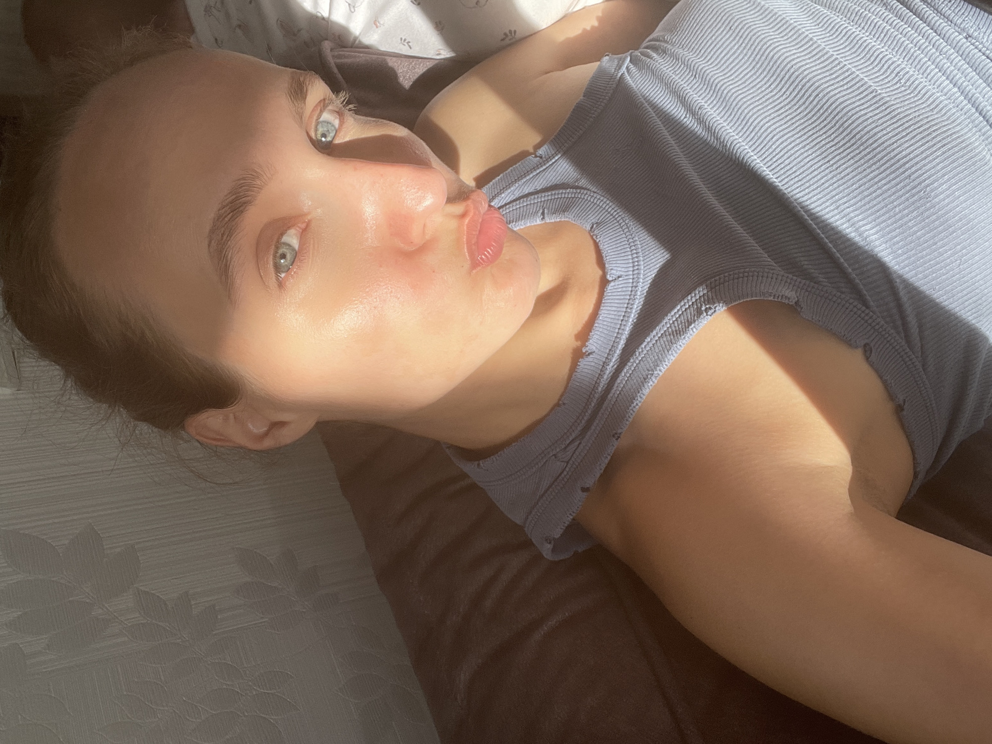 An individual, appearing to be a young woman, is captured in a close-up, diagonal shot, likely a selfie, indoors in Sharkan, Russia. She is lying down, looking directly at the camera with bright blue-grey eyes and a playful pouting or "kissing" expression. Her light brown hair is pulled back, revealing her forehead. Her skin, particularly on her forehead and cheeks, reflects strong, warm natural sunlight, giving it a dewy or shiny appearance.
She is dressed in a light blue or grey ribbed tank top, the texture of which is clearly visible. The sunlight streams in from the upper left, casting distinct shadows across her body and face, creating a strong contrast between illuminated and shaded areas. Behind her head, prominent shadows resembling leaves are cast upon a light-colored surface, possibly a pillow or wall, suggesting foliage outside a window. She rests on a dark brown surface, possibly a bed or sofa. The overall scene conveys a relaxed daytime moment, illuminated by abundant natural light. There is no visible text in the image.