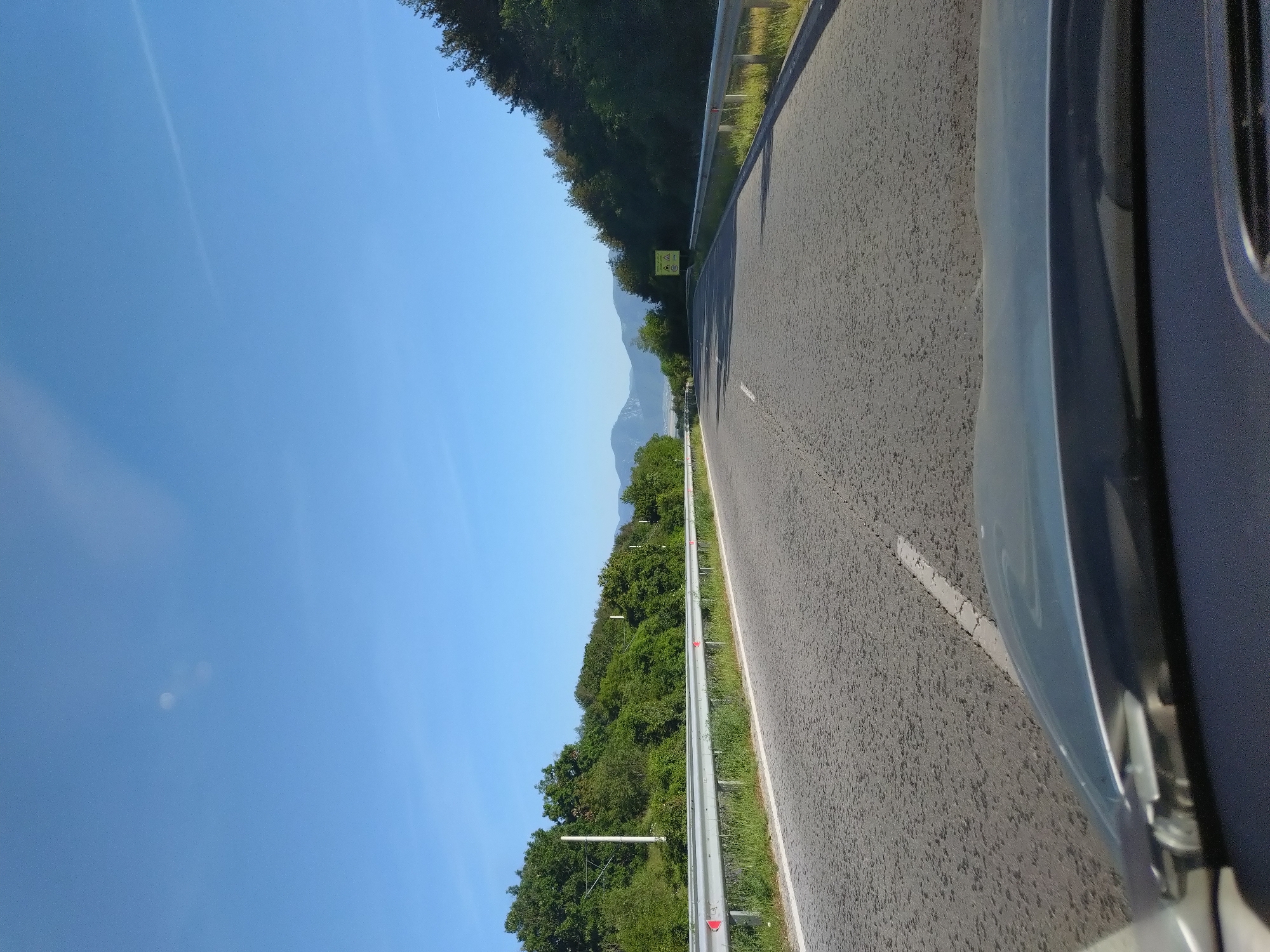 The image depicts a view from the perspective of a driver on a paved road. The road has a white dashed line marking the lane. On either side of the road, there are metal guardrails and lush green trees. In the distance, mountains are visible under a clear blue sky with a few wispy clouds. The scene appears to be a highway in a rural or semi-rural area, possibly near Zmeyovo, Bulgaria, given the characteristic landscape. There are no people visible in the frame.