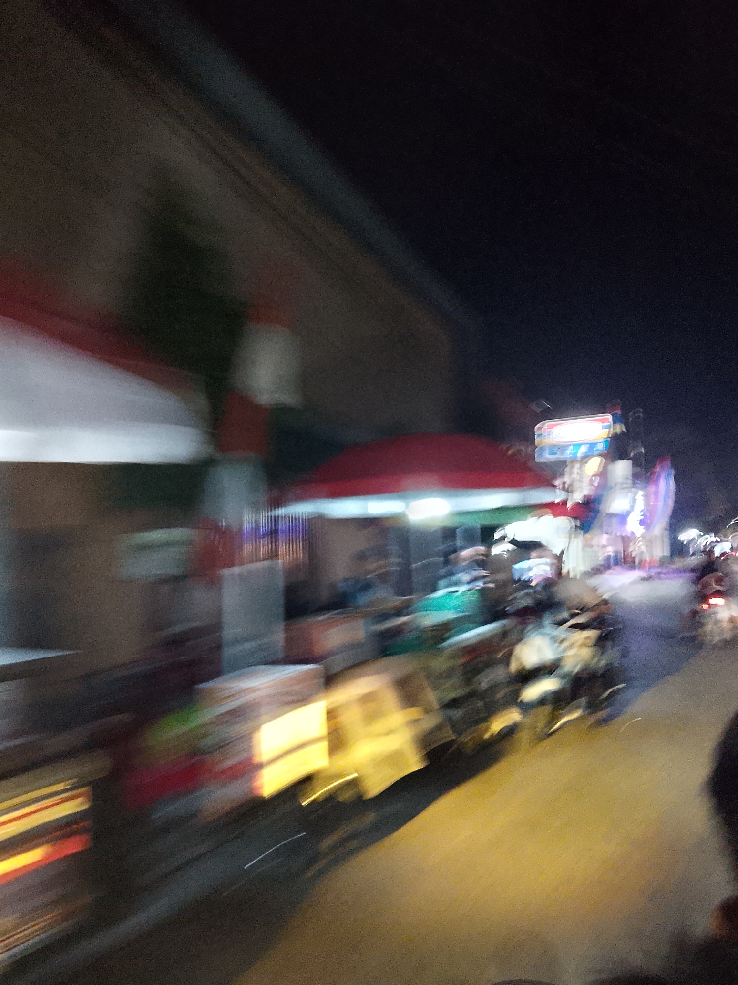 The image depicts a blurry, motion-filled night scene on a street in Ngelo, Indonesia. The scene appears to be a commercial area with several stalls and shops lining the road. A red canopy structure is visible on the left, with bright lights illuminating the area underneath. Several people are present, though their features are indistinct due to motion blur. They appear to be interacting at the stalls, possibly as vendors or customers.
A sign with what looks like blue and yellow lettering is prominently displayed on a pole in the background. Several motorcycles are also visible, some parked and others in motion, contributing to the sense of activity. The street is illuminated by artificial lights, casting a warm yellow glow on the road surface. The overall impression is a lively night market or street scene, capturing the dynamic atmosphere of a busy Indonesian street at night.