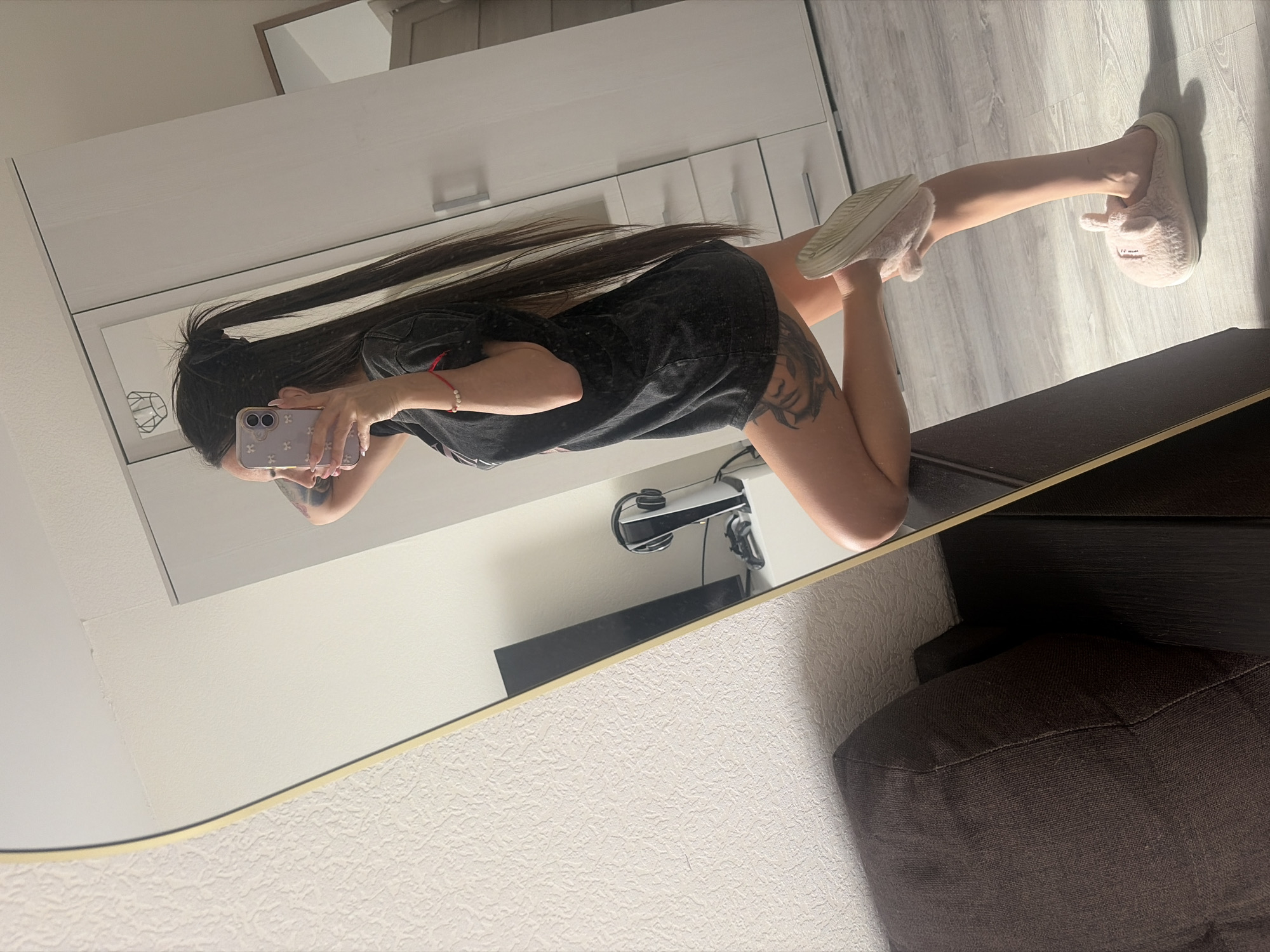 A person is taking a mirror selfie in an indoor setting. The person is wearing a dark t-shirt and has long, dark hair. They are holding a phone with a light-colored case in their hand. The person is wearing light-colored slippers on both feet, one of which is raised. The room appears to be a bedroom, with a white dresser in the background and a dark brown couch visible in the foreground. A desk lamp is also visible on top of the dresser. The wall is light-colored with a textured pattern. The city and exact location in Saint Petersburg, Russia cannot be determined.
