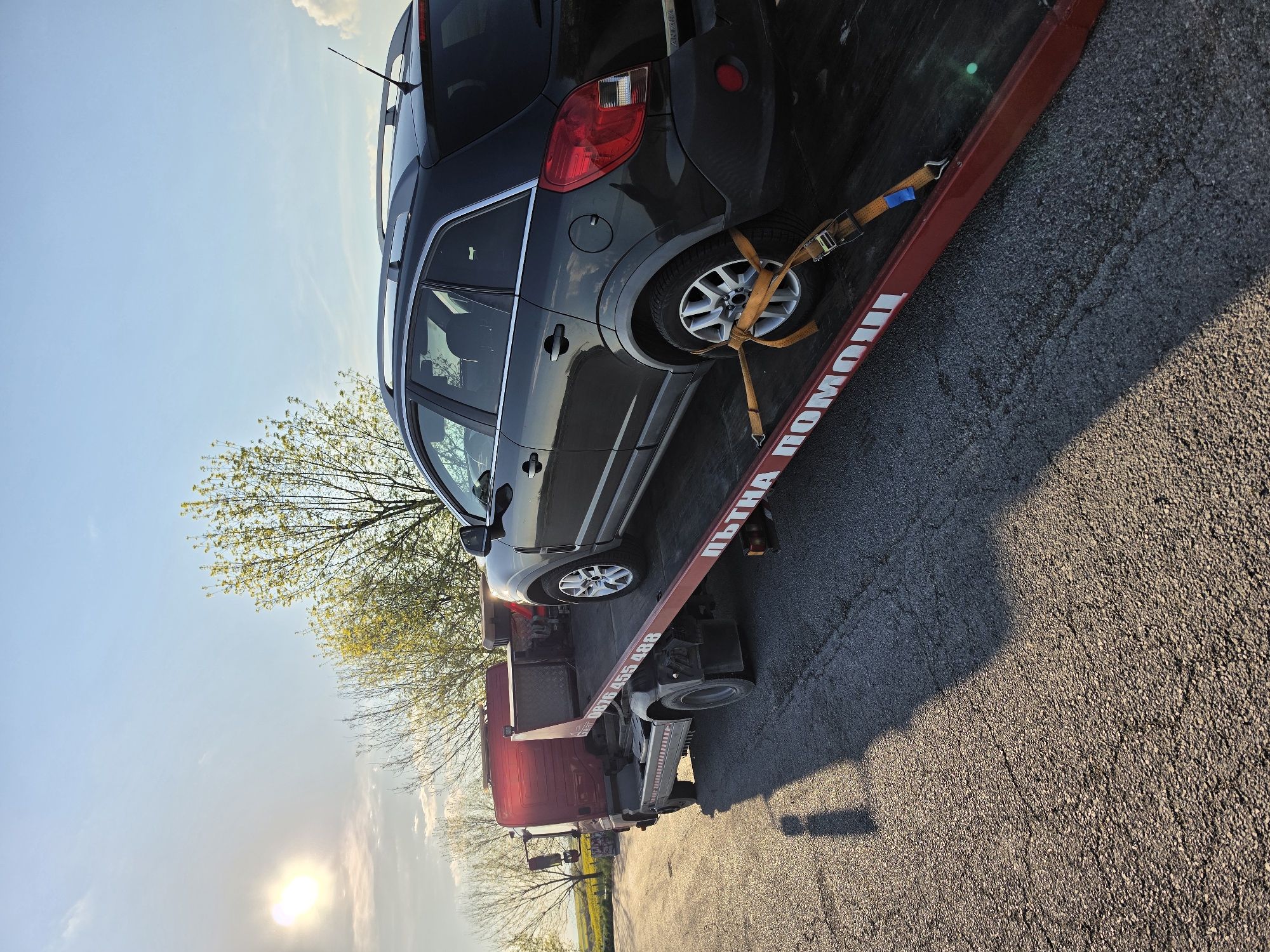 A dark grey Opel Antara SUV is secured on the back of a red tow truck with yellow straps attached to its rear driver's side wheel. The tow truck has white lettering in Bulgarian ("ПЪТНА ПОМОЩ") along the side of its loading platform. The scene is set on an asphalt road, with a field and trees visible in the background. The sun is low in the sky, creating long shadows and a bright glare. The tow truck may be located in Polsko Padarevo, Bulgaria, based on information included in the prompt.