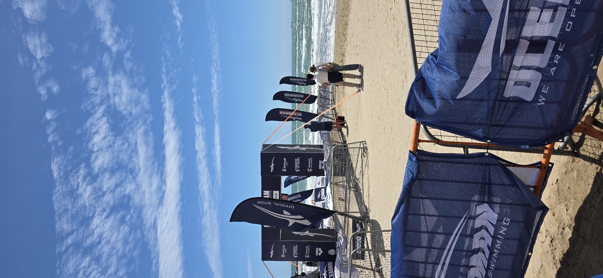 Here is a description of the image:
The image captures a beach scene in Benicàssim, Spain, with a view of the ocean on the horizon. The sky is clear with some wispy clouds.
In the foreground, there are barriers and flags set up. The flags are dark blue and display the logos of several entities, including "OCEANMAN", "Benicassim", "arena", and "Diputació de Castelló". The banners have an orange frame. Several people are standing on the beach further away in a huddle, their backs to the camera, seeming to look out towards the sea. The sand is light brown. The surf is relatively calm with small waves visible.
