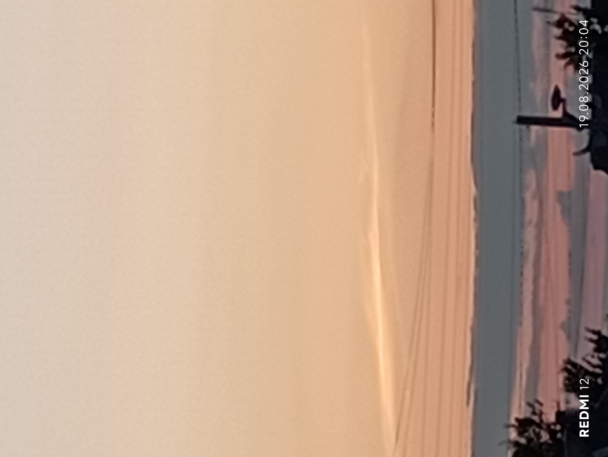 The image displays a dusky sky with hues of orange and pink, indicative of either sunrise or sunset. A thin, wispy cloud formation is visible horizontally across the upper-middle section of the frame. Below this, power lines are stretched horizontally, partially obscured by the ambient light. The lower portion of the image shows a silhouetted landscape, with the dark outlines of trees and structures against the softer colors of the sky. In the bottom left corner, text reads "REDMI 12". In the bottom right corner, text reads "19.08.2026 20:04". There are no identifiable people or specific landmarks that would allow for a precise location within the city of Smila, Ukraine. The scene is a general depiction of a sky at twilight with some urban or suburban elements in the background.