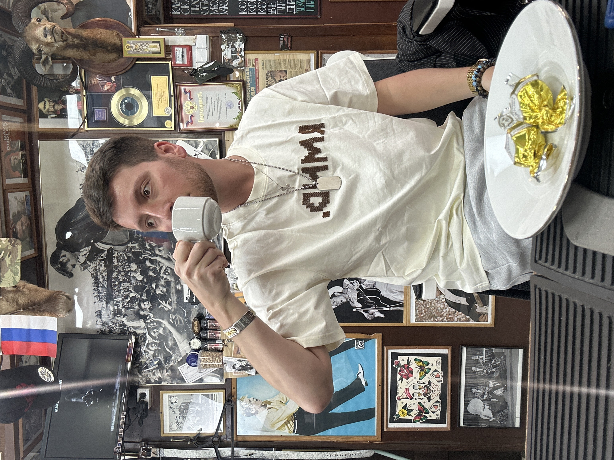 The image presents a Caucasian man holding a white cup and saucer, possibly drinking or about to drink. He is seated in a room densely decorated with framed pictures, certificates, taxidermied animals, and various objects adorning the walls. The overall arrangement suggests a somewhat cluttered and personalized interior.
The man is wearing a light-colored T-shirt with the word "ЯЩИНО" written across it and a silver chain with a cross and dog tags. He is also wearing a wristwatch and multiple bracelets. The saucer he holds has a golden decoration.
A Russian flag is visible, along with Cyrillic writing on some of the objects and signs, confirming a location in Russia. The room's collection of memorabilia implies a sense of history and personal significance, possibly a barbershop, given the scissors and grooming supplies visible. Therefore, the location is in Moscow, Russia.