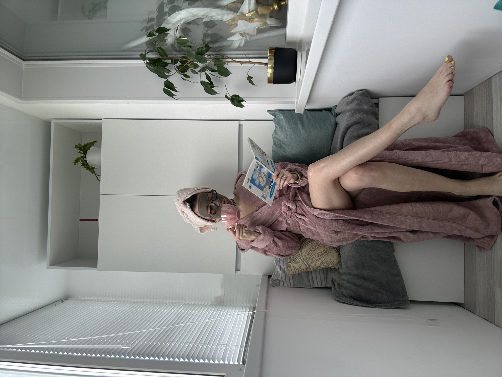A woman with a pink towel on her head and a face mask sits on a bench with her legs crossed, holding a pink teacup in her left hand and an open book in her right hand. She is wearing a pink robe and glasses. The scene takes place on a narrow balcony, with white blinds on the left and a window on the right. A potted plant sits on the windowsill. There is a white cabinet with shelves in the background. The floor is grey.