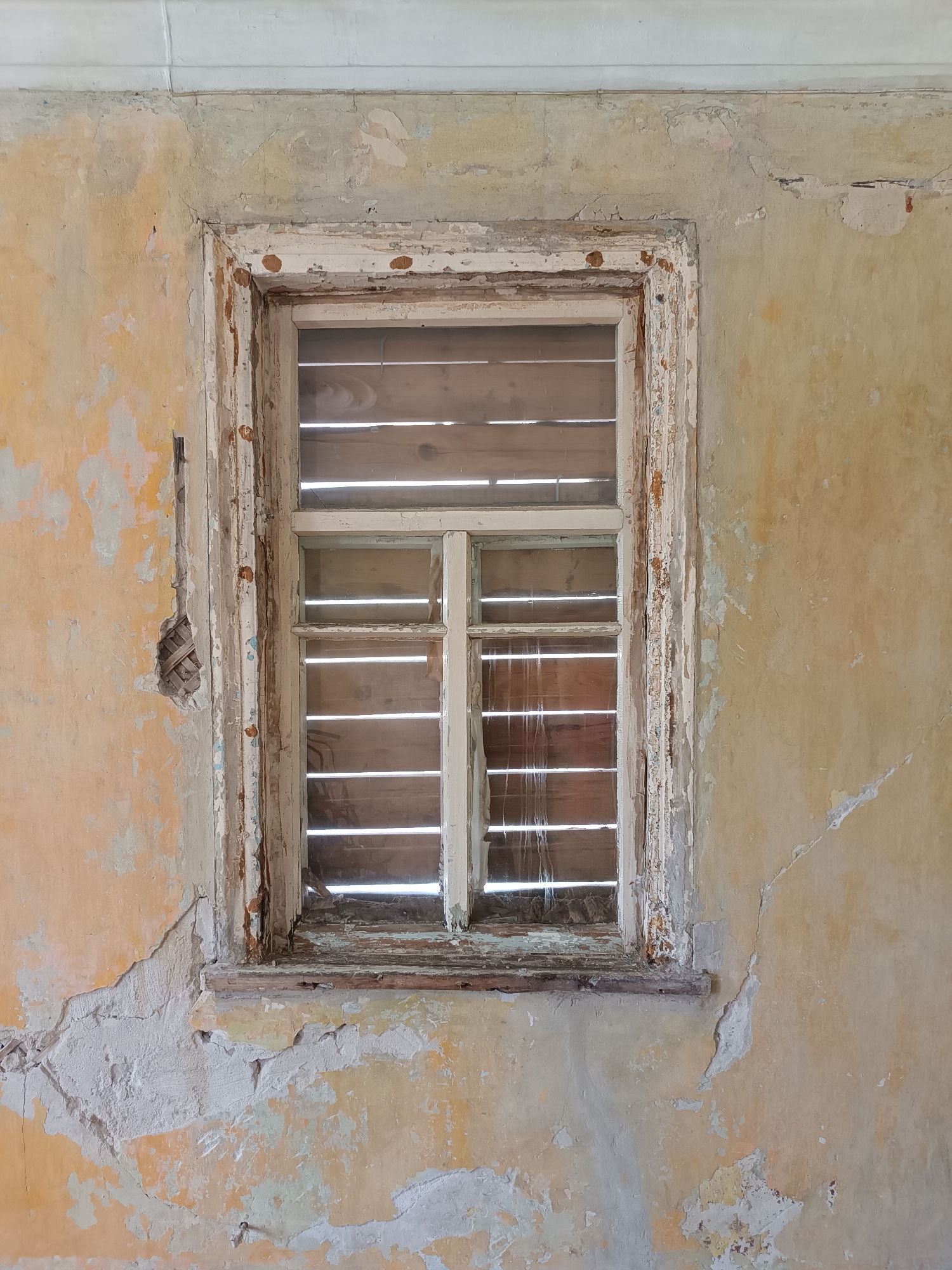 An abandoned window in a dilapidated building, Yelets, Russia. The window is set within a peeling, textured wall, predominantly yellow with patches of white and grey plaster. The window frame itself is weathered and splintered, with visible nails. The window pane is covered with horizontal wooden slats, obscuring the view beyond. Light filters through the gaps in the slats, creating horizontal lines of illumination. The wall exhibits significant wear and tear, with large areas of plaster missing, revealing underlying textures and structural elements.
