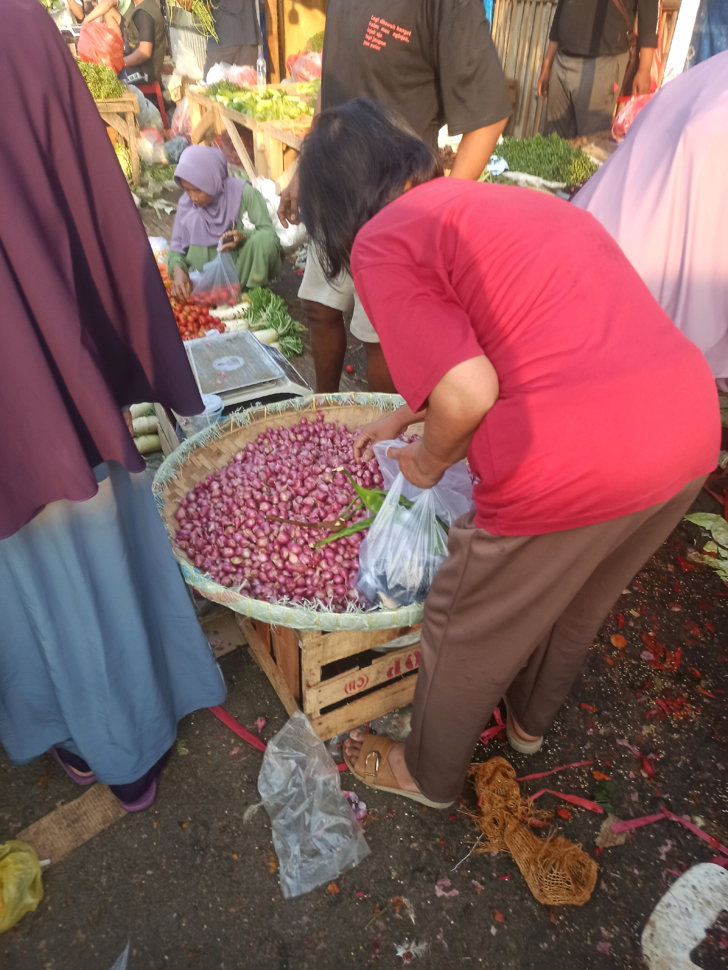 A vendor, wearing a red shirt and brown pants, is bagging red onions at a market in Cikarang, Indonesia. She is standing next to a large woven basket filled with red onions. Another woman, wearing a purple hijab and green dress, is kneeling on the ground to her left, also surrounded by produce. In the background, other people and stalls are visible, suggesting a busy marketplace. The ground is paved and appears somewhat dirty, with scattered debris and petals.