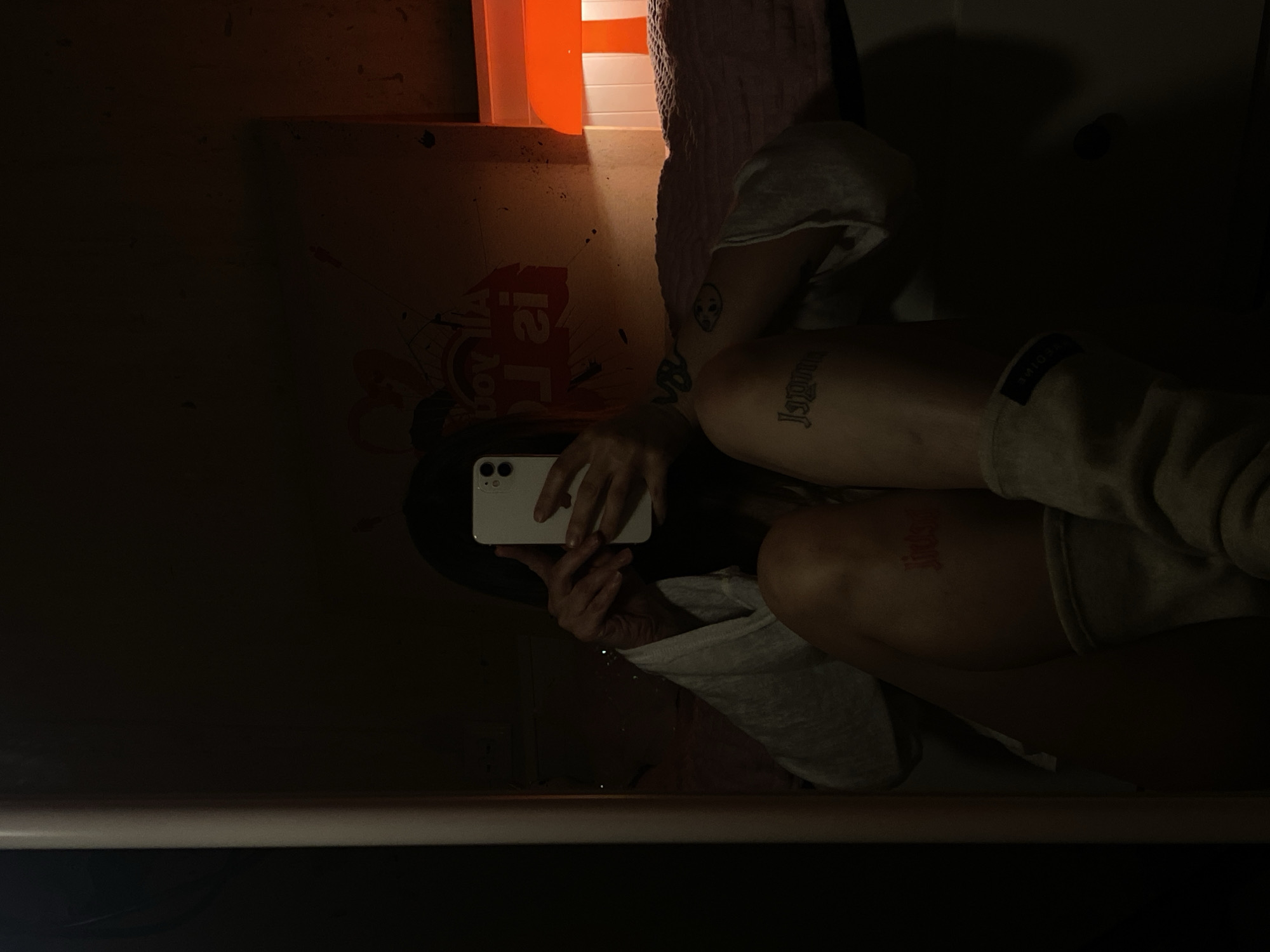 A person is reflected in a mirror, holding a white smartphone and taking a picture of themselves. The background includes an orange bin, a pink towel, and a wall with faded graffiti and logos, including what appears to be an orange Home Depot logo. The lighting is dim. There is no information available about the location in the city of Votkinsk, Russia.