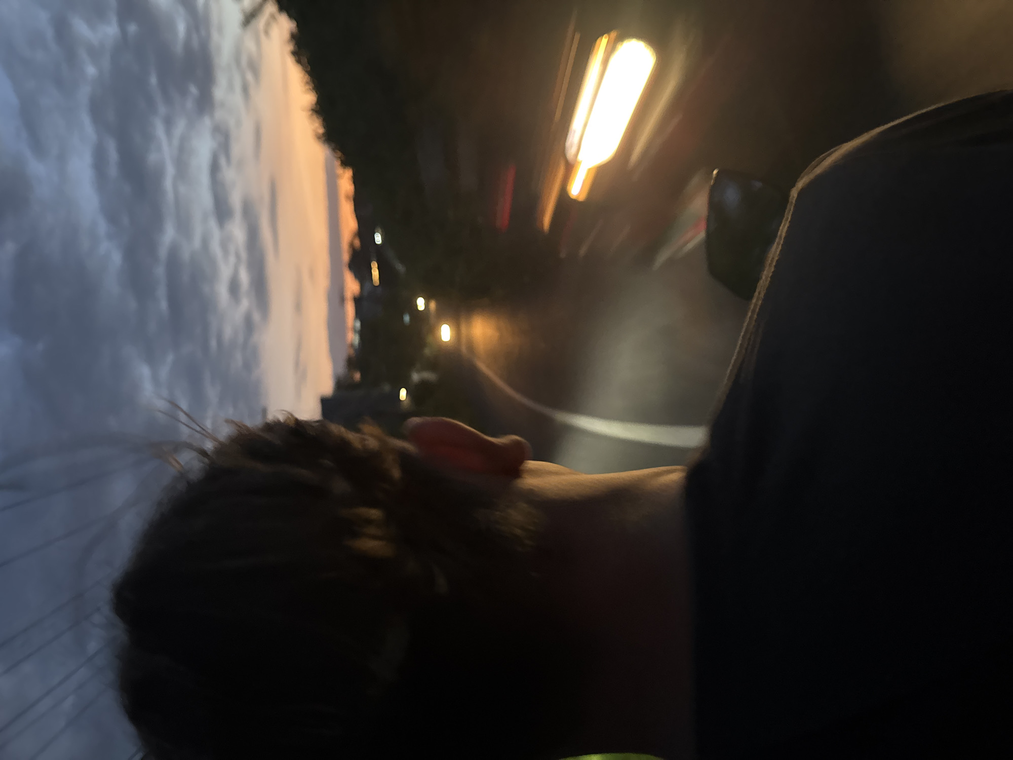 The image shows a person with dark hair and a visible ear riding a vehicle on a road at dusk in what is possibly Kuta Selatan, Indonesia. The sky on the left exhibits dark, cloudy formations transitioning into orange hues. On the right, there is a road with blurred street lights, suggesting motion and speed. The overall environment is dimly lit. The person appears to be in motion, with a blurry foreground, giving a point-of-view perspective from the vehicle they are riding.