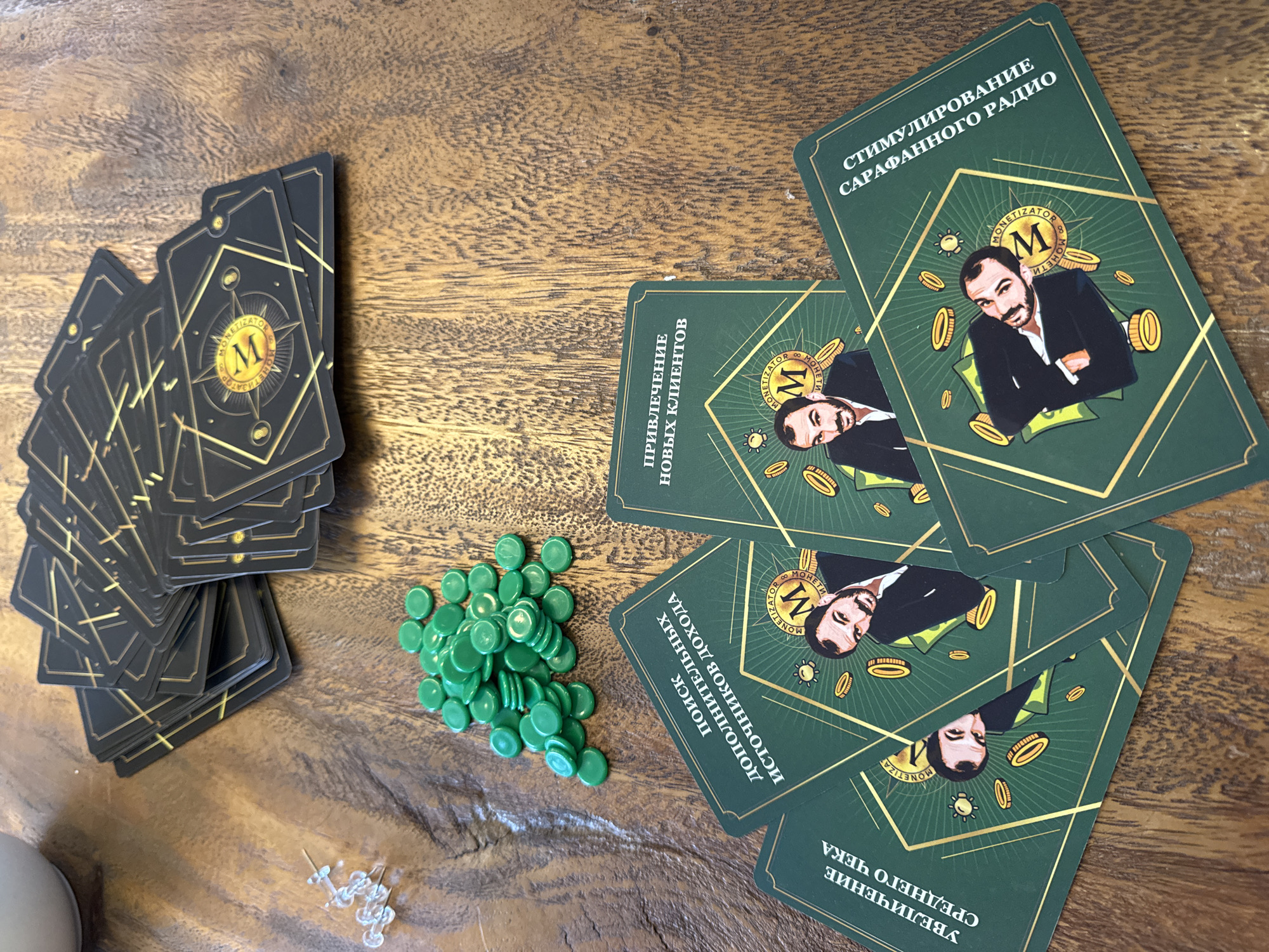 This high-angle shot displays components of what appears to be a board or card game laid out on a warm-toned wooden surface with a visible grain. The scene was captured in North Kuta, Indonesia.
On the left, a large stack of dark-colored cards is present, their backs featuring a gold geometric pattern with a central compass-like emblem enclosing a stylized 'M' and the word "MONETIZATOR". To the right of these, a pile of small, round, flat green tokens or chips is gathered.
On the right side of the frame, several green cards are fanned out, revealing their fronts. Each card features an illustration of a bearded man in a suit, surrounded by floating gold coins and banknotes, with a similar 'M' and "MONETIZATOR" logo. Visible text on these cards, written in Cyrillic, indicates various business or marketing strategies, suggesting the game's theme. The texts include: "СТИМУЛИРОВАНИЕ САРАФАННОГО РАДИО" (STIMULATING WORD-OF-MOUTH RADIO), "ПРИВЛЕЧЕНИЕ НОВЫХ КЛИЕНТОВ" (ATTRACTING NEW CLIENTS), "ПОИСК ДОПОЛНИТЕЛЬНЫХ ИСТОЧНИКОВ ДОХОДА" (SEARCH FOR ADDITIONAL SOURCES OF INCOME), and "УВЕЛИЧЕНИЕ СРЕДНЕГО ЧЕКА" (INCREASING THE AVERAGE CHECK/BILL).
In the bottom left corner, a small cluster of clear plastic thumbtacks or pushpins is visible, alongside a partial view of a white object, possibly a cup or container. The overall arrangement suggests a game in progress or set up for play.