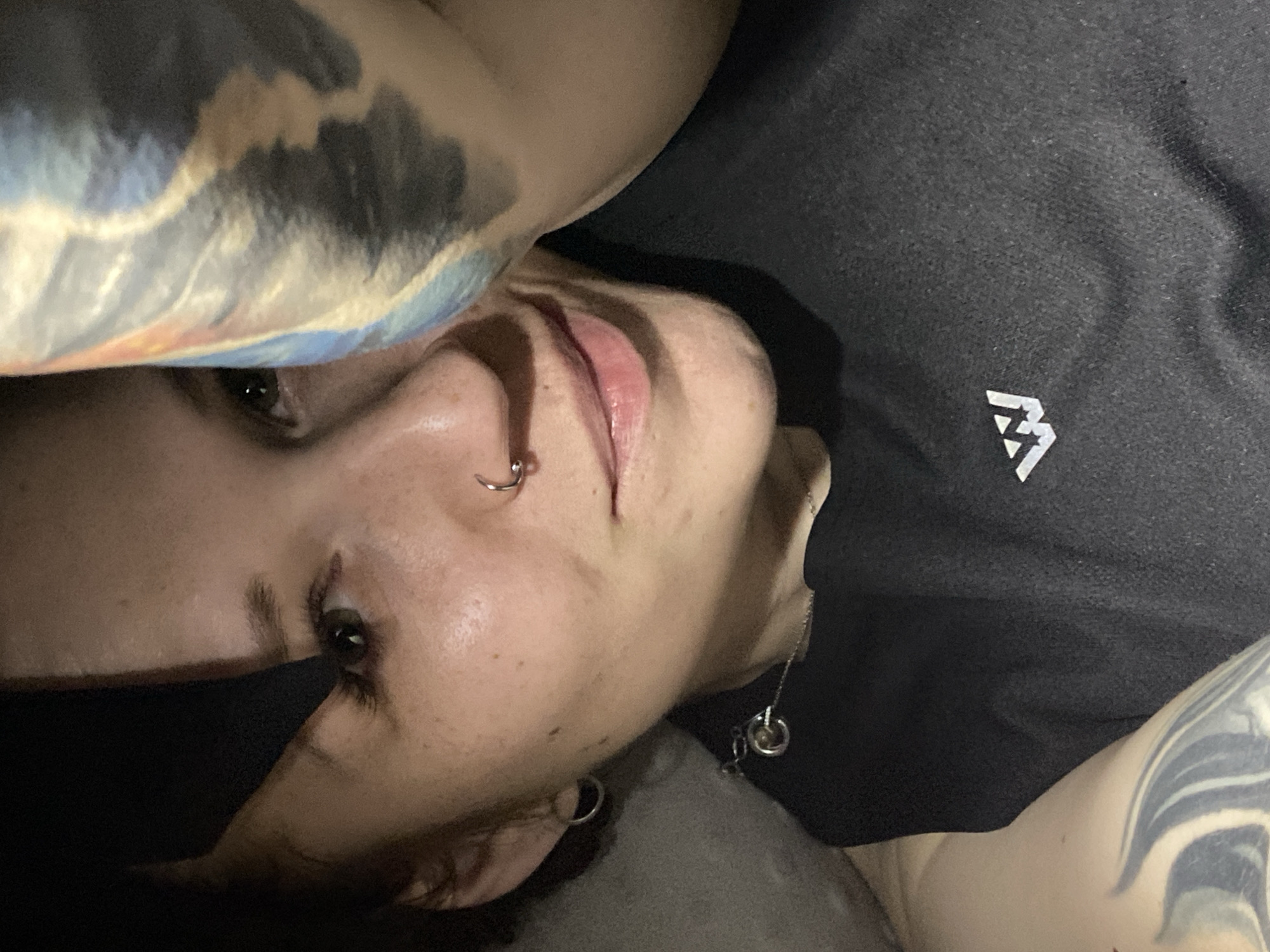 A selfie-style photograph depicts a person with fair skin, lying down and looking up at the camera at an angle. The person has dark hair, visible eyelashes, and a piercing in their left nostril. They are wearing a black top with a white logo on the right chest area. A silver necklace with round pendants is visible around their neck. Tattoos are present on both arms, with a design featuring dark and light shades of blue, grey, and white. The background is out of focus, but suggests a dark, possibly indoor environment. The image does not contain discernible information about the city of Chaikovsky, Russia.