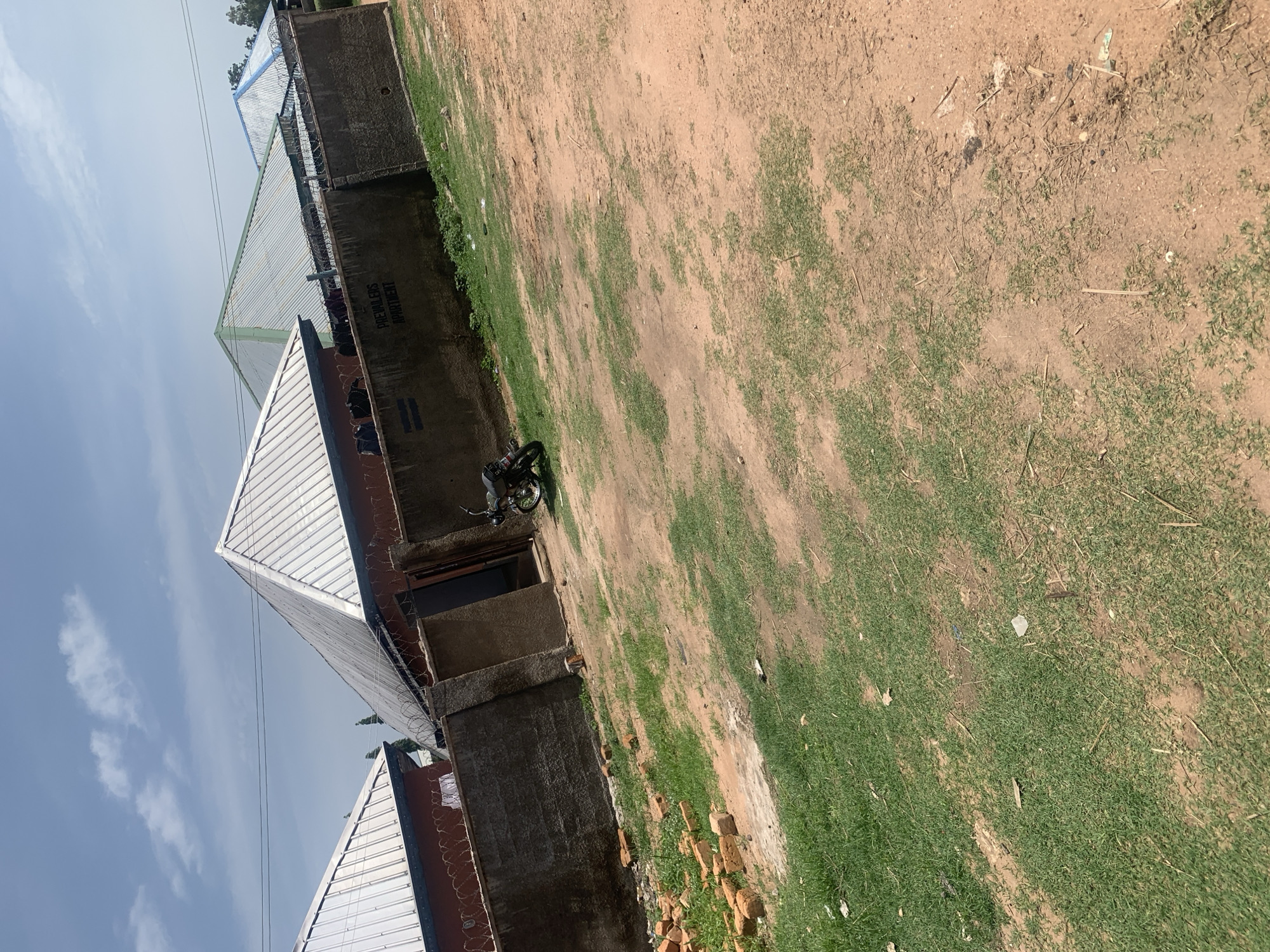 The image depicts the exterior of "PREVALERS APARTMENT" in Jauro Yinu, Nigeria. The property is enclosed by a concrete wall topped with barbed wire. Several buildings with corrugated metal roofs are visible behind the wall. A motorcycle is parked on the unpaved ground in front of an open doorway. The ground is a mix of dirt and patchy grass. A pile of bricks lies near the wall in the foreground. The sky is blue with scattered clouds.