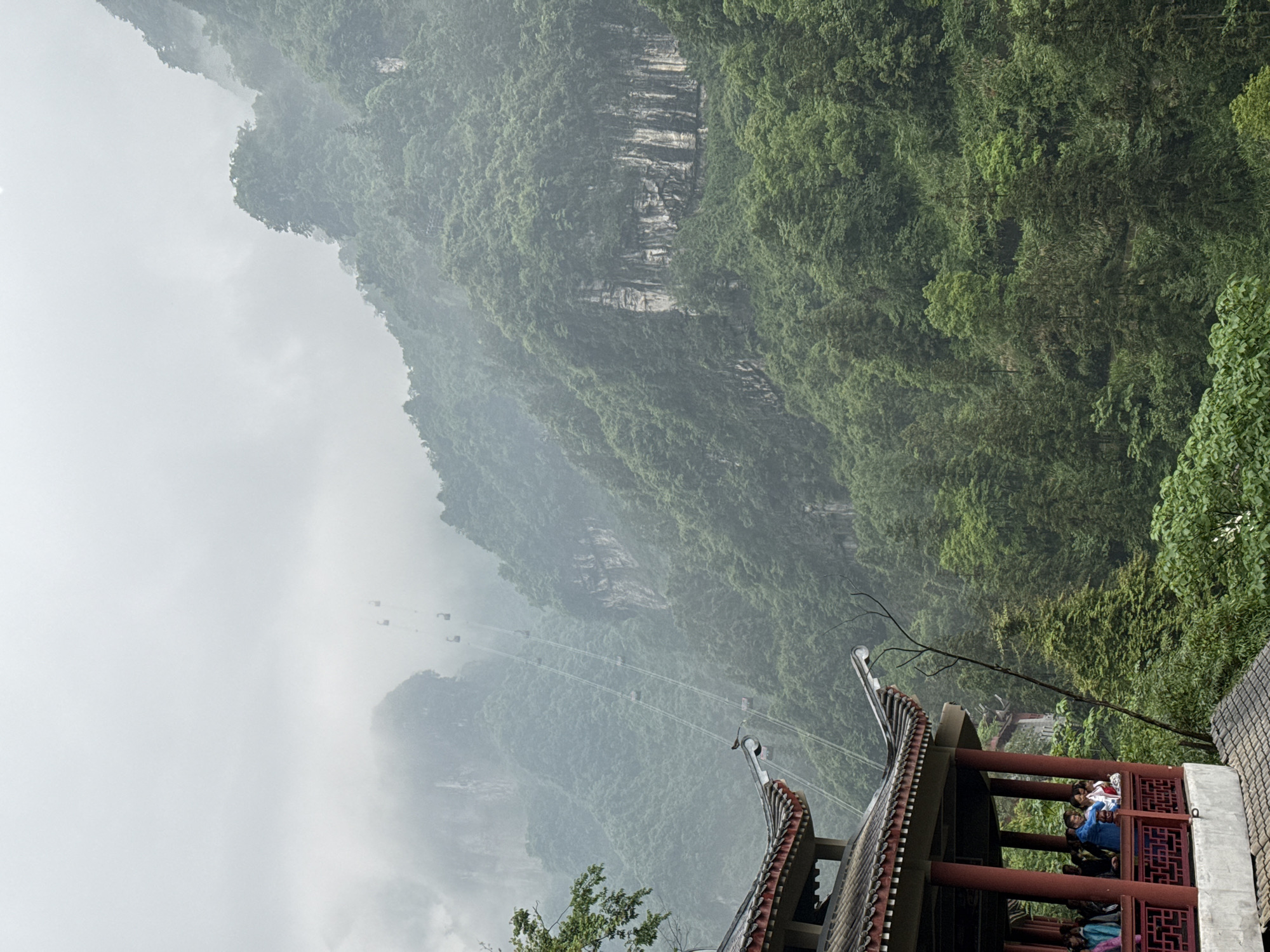 An eye-level shot depicts a mountain range in Yongding District, China, partially obscured by fog and lush with green trees. A cable car system, with visible cables and cars, spans across the mountains. A traditional Chinese building is in the foreground, with a tiled roof and decorative red latticework. People are gathered on the building’s platform, some wearing blue shirts and looking out at the view.