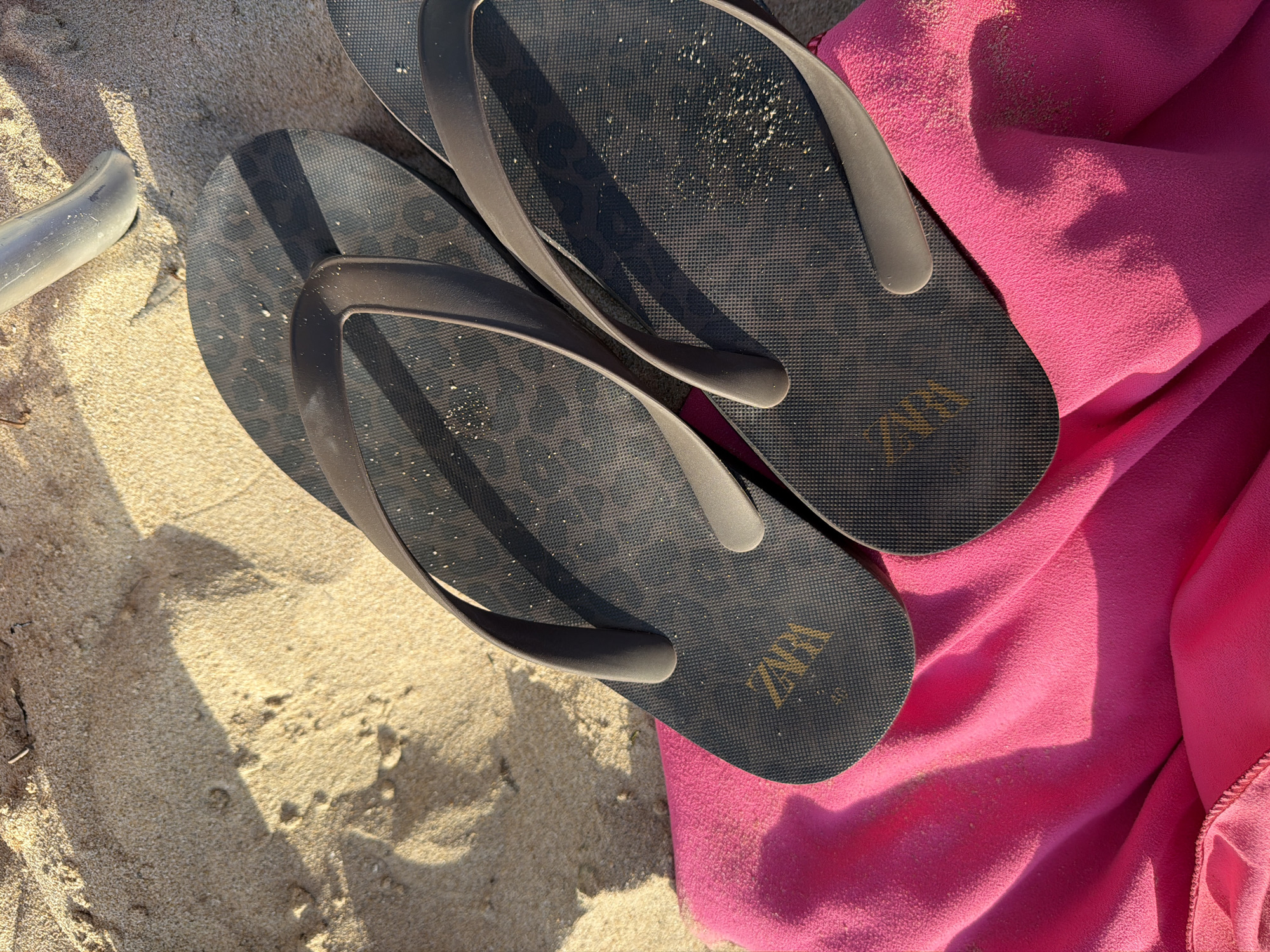 Two dark gray flip-flops with a subtle leopard print pattern are laid on a bright pink beach towel. The flip-flops are positioned on a sandy surface, with the towel beneath them. One flip-flop is positioned slightly above and to the left of the other. The right flip-flop has "ZARA" imprinted in gold lettering on its sole. Sand particles are visible on the surface of the flip-flops and the towel. In the background, a portion of a light-colored plastic object, possibly a chair leg or a similar beach accessory, is visible on the sand. The lighting suggests a sunny day, with distinct shadows cast by the flip-flops and the plastic object. The scene appears to be set on a beach in Urbanització Cales I Talaies, Spain.