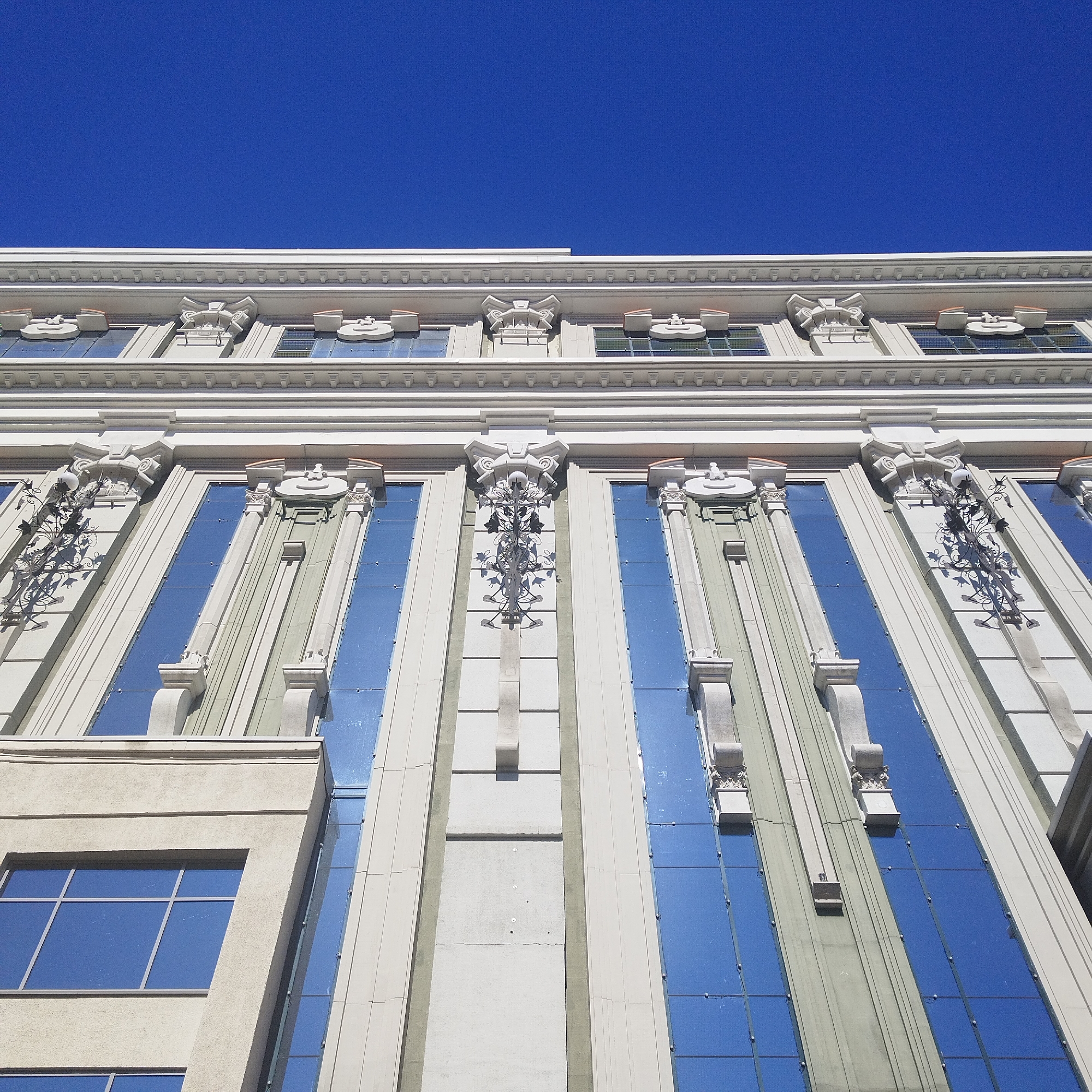 This image captures a low-angle, upward view of a building facade in Yekaterinburg, Russia, set against a pristine, vibrant blue sky. No people or activities are visible.
The architecture suggests a classical or Art Deco influence, characterized by its symmetrical design, ornate detailing, and strong vertical lines. The building's primary structure is a light, possibly white or beige, material, providing a sharp contrast with the expansive blue sky and the reflective, blue-tinted windows.
Prominent architectural features include vertical columns or pilasters adorned with elaborate, stylized capitals and decorative scrollwork. These elements frame tall, narrow windows, many of which strongly reflect the blue sky, while others show internal grids. Between some of the window sections, there are areas with a greenish-grey hue, adding subtle color variation.
Intricate decorative motifs are present along the building's cornices and on the upper parts of the columns, including sculpted forms and projections that create visual depth. On one of the left vertical sections, an ornamental ironwork fixture, possibly a historical lampholder, adds a touch of intricate design.
The scene is brightly lit by direct sunlight, casting distinct shadows that accentuate the building's three-dimensional details. This strong illumination, combined with the clear, cloudless sky, indicates a sunny day. The perspective emphasizes the building's height and detailed craftsmanship. No text is visible anywhere in the image.