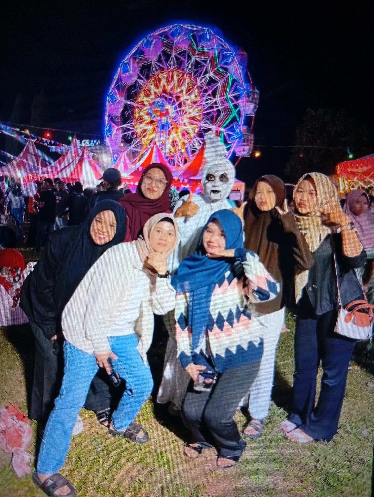 A group of seven people are posing for a photo at night with a Ferris wheel in the background. The Ferris wheel is illuminated with colorful lights, casting a vibrant glow on the scene. The group includes six women and one person dressed in a white costume with black face paint, resembling a ghost.
The women are dressed in various outfits, most of them wearing hijabs. One woman in the front left is wearing a black hijab and a white sweater over light blue jeans. Next to her, another woman in a white sweater and light blue jeans is posing with her chin resting on her hand. To their right, a woman in a blue hijab is smiling and holding her hand to her cheek. Behind them, a woman in a maroon hijab is smiling and wearing glasses. Another woman in a brown hijab is making a peace sign. The woman on the far right, also in a hijab, is wearing a dark jacket and holding a small white purse.
The person in the ghost costume is standing in the back, with their mouth open and eyes painted black. The ground appears to be covered in grass, and there are tents and other structures in the background, suggesting a fair or festival. The time of day is clearly night due to the dark sky and the illuminated attractions. The overall mood seems festive and joyful, with several people smiling for the camera. The visible text in the background reads "LGCRAY". The location context is Lhokseumawe, Indonesia.
