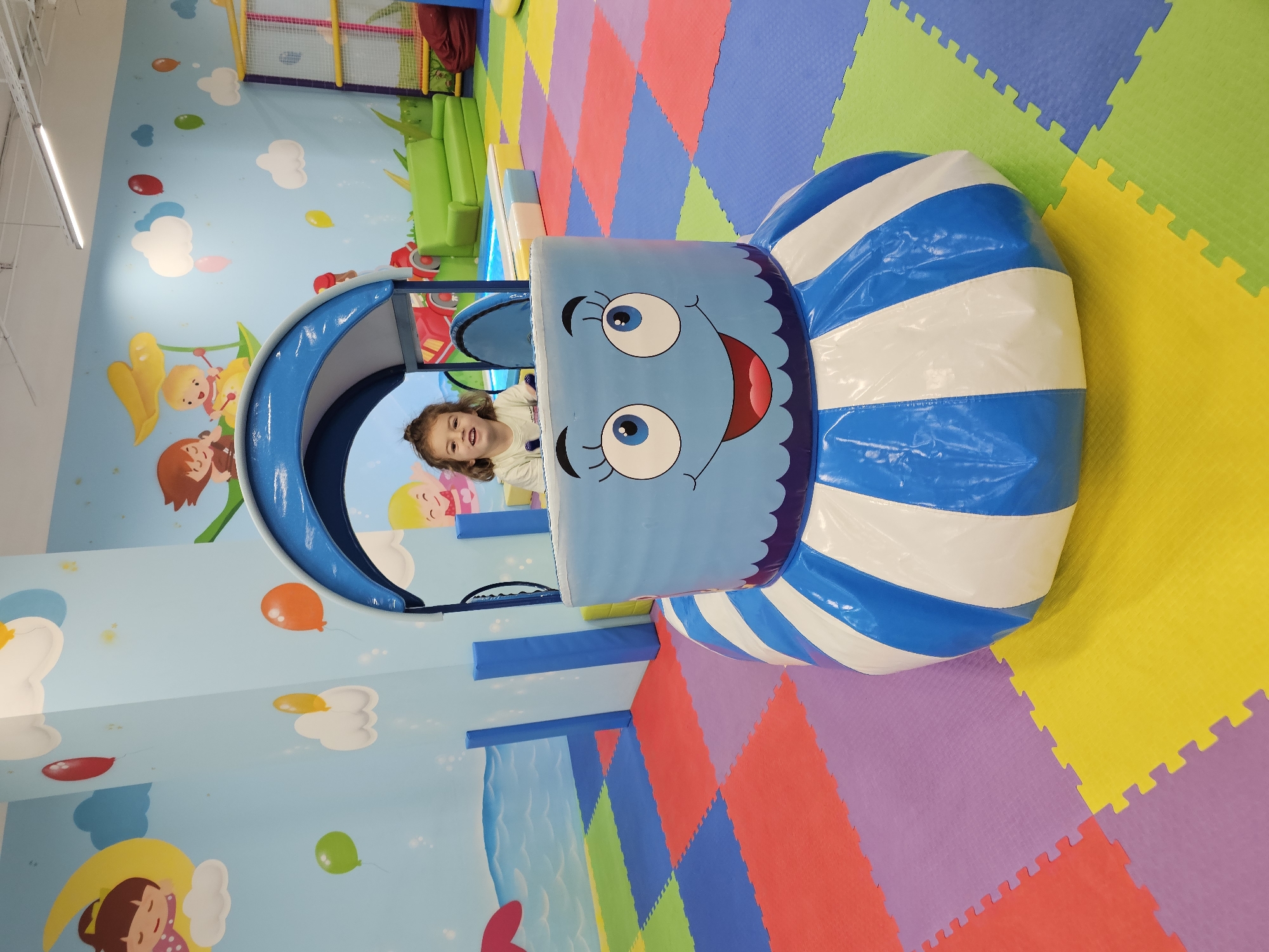 A slightly high-angle, indoor shot of a playground. A young girl is seated inside a blue and white striped play structure with a cartoon face on the front. The playground has colorful interlocking floor mats, a mural on the wall, and various other play structures in the background. The mural features cartoon characters, clouds, and balloons. The location appears to be indoors. The city of Moscow, Russia is not discernible.