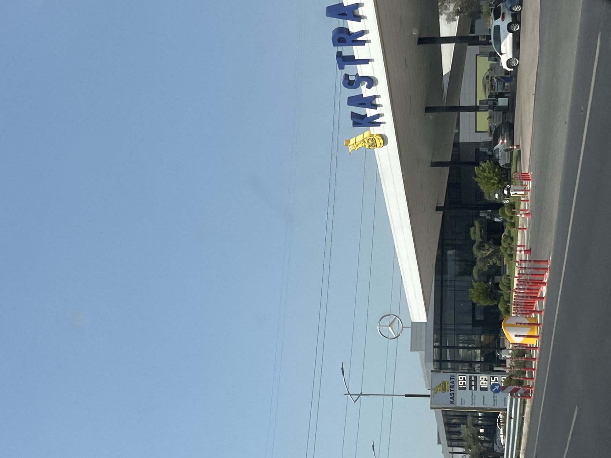 The image shows a view of a modern gas station with a large canopy in Katund i Ri, Albania. The KASTRATI logo is prominently displayed in large blue letters on the side of the canopy, accompanied by a yellow graphic. To the left of the gas station, a Mercedes-Benz dealership is visible, with a large Mercedes-Benz logo displayed on the building. A price board at the gas station shows fuel prices, with "Diesel" listed at 1.99, and two other fuel types at 1.89 and 0.75. Several cars are parked at the gas pumps under the canopy, and people are visible tending to the pumps. The scene is set against a clear blue sky. The foreground shows a road with safety cones and barriers indicating a designated area.