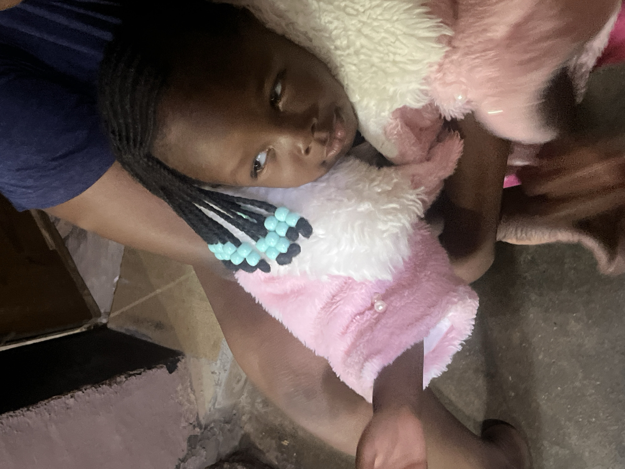 A close-up, slightly tilted portrait captures a young child, likely female, with dark skin and braided hair adorned with light blue and black beads. The child is resting or reclining, looking upward and to her right with a neutral or contemplative expression. She is partially enveloped in a fluffy, pink and white garment or blanket, possibly a hooded jacket or plush item, which features a small, pearl-like button. A dark blue fabric, potentially part of another person's clothing or a cushion, is visible near the child's left side. Parts of what appear to be an adult's arm or leg are visible, supporting or holding the child. The background reveals rough, dark, and textured surfaces, consistent with concrete or unfinished walls and flooring, suggesting an indoor or sheltered setting. The scene takes place in Gura Top, Nigeria. No text is visible in the image.