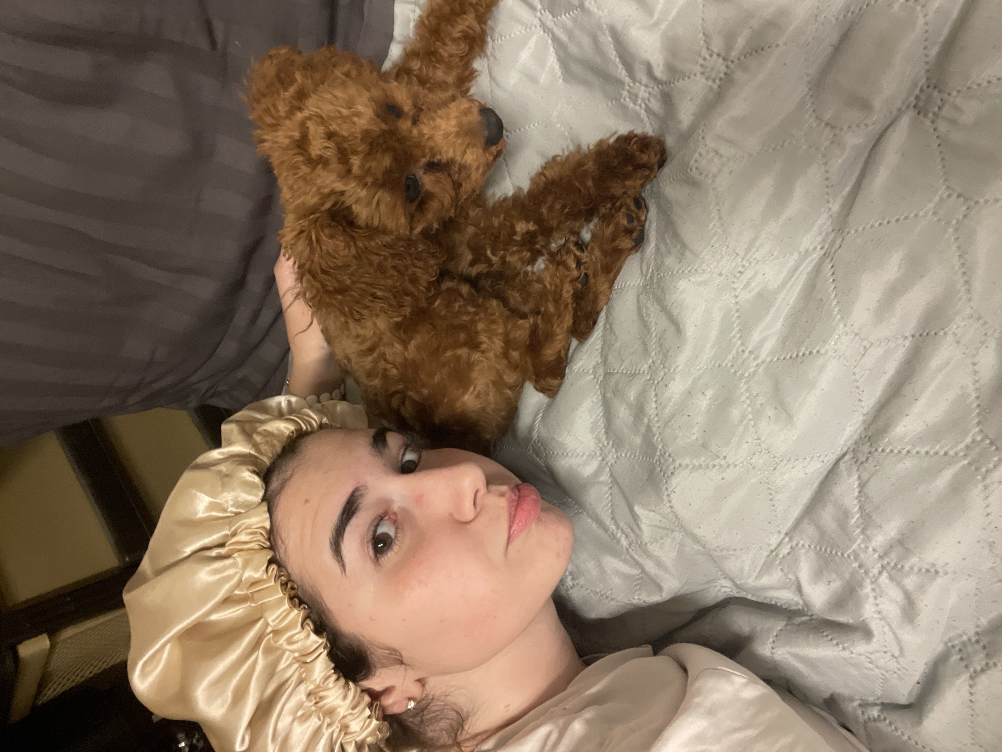 A young woman in a gold satin bonnet lies on a light gray, patterned quilt. She looks up at the camera with a slight pout. Above her, a brown plush teddy bear lies on the quilt, partially held by the woman's left hand. The environment includes a dark gray blanket on the left and a glimpse of a dark brown cabinet. The image lacks sufficient information to determine its location in Sofia, Bulgaria.