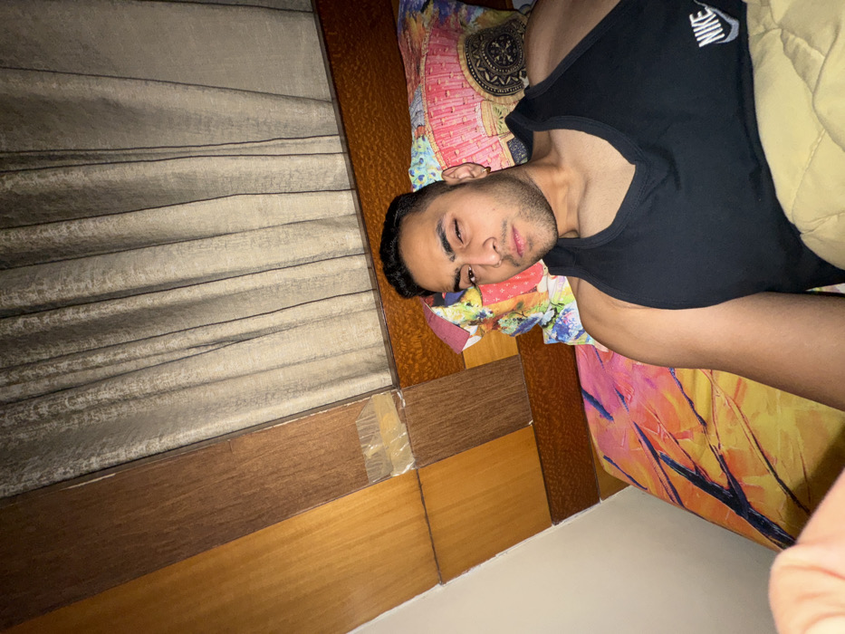 A man in a black tank top lies on a colorful patterned bed with a tan blanket. He appears to be sleeping or resting, with eyes closed. The scene is a bedroom with curtains and a wooden-paneled wall. Some tape is visible on the wall. The image is taken at a slight angle. A Nike logo is on the tank top.