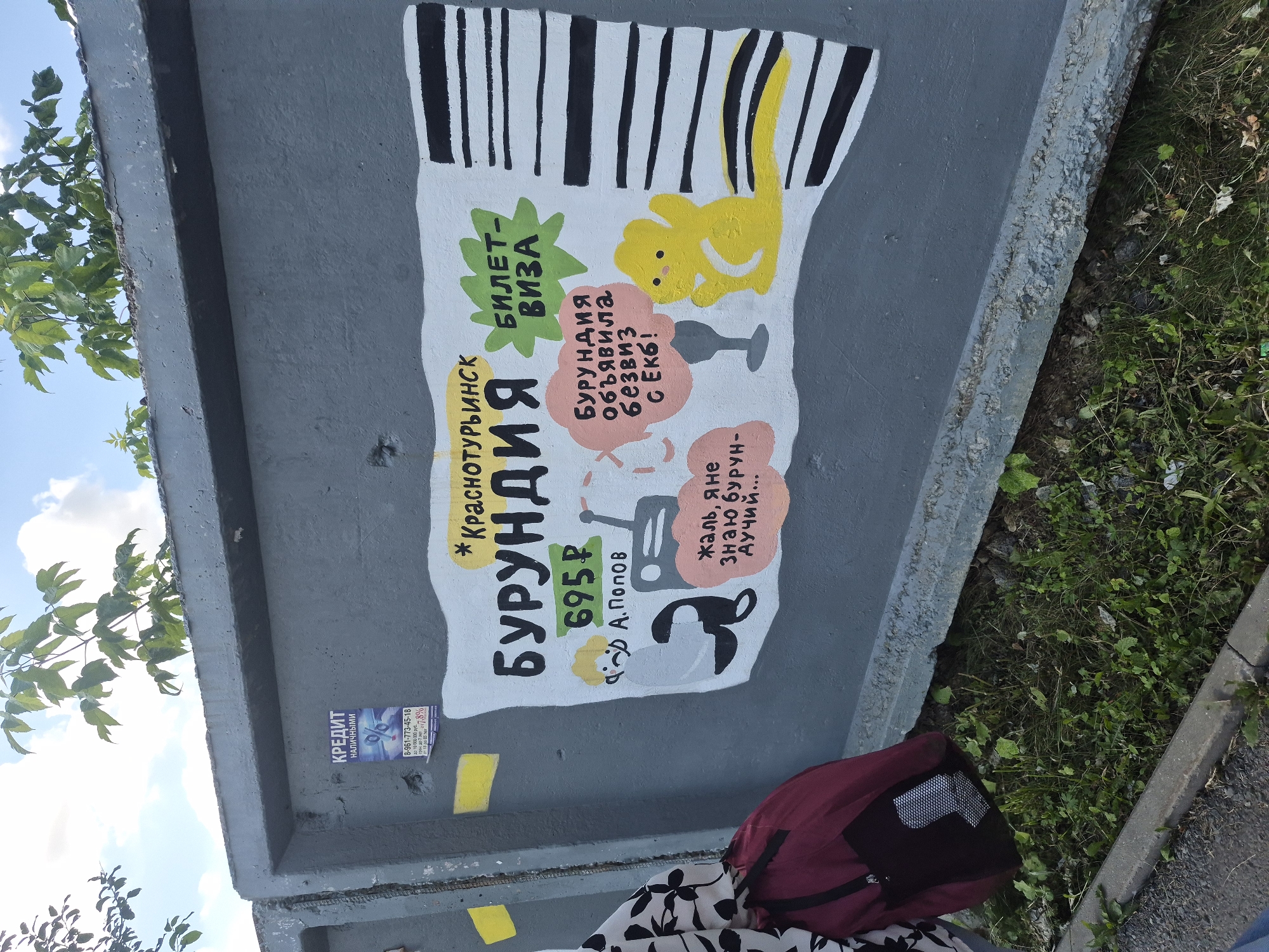 The image depicts a mural painted on a concrete wall in Yekaterinburg, Russia. The mural is a colorful, cartoonish advertisement with a ticket motif. Dominating the scene is the word "БУРУНДИЯ" (Burundia) in large, bold letters, with "БИЛЕТ-ВИЗА" (Ticket-Visa) written in a green starburst above and to the right. Below "БУРУНДИЯ" is a price of "695 P" (695 rubles). To the left of the price, a cartoon figure of a person with yellow hair and a sad expression is depicted. To the right of the price, a gray radio and a pink speech bubble containing the text "Бурундля объявила безвиз с ЕКБ!" (Burundia announced visa-free travel to EKB!) are shown. A small, yellow, cartoon animal with large eyes and paws is located to the right of the speech bubble. Below the main text, a smaller speech bubble contains the text "Жаль, я не знаю бурун-дучий..." (Too bad, I don't know Burundian-Duchy...). The background of the mural resembles a barcode with black and white stripes.
On the upper left portion of the concrete wall, a small flyer or poster is affixed, advertising "КРЕДИТ НАЛИЧНЫМИ" (Cash Loan) with contact information. Green foliage from trees hangs over the top of the concrete structure.
In the foreground, a person's torso and shoulder, wearing a black and white patterned garment, are visible on the left side of the image. A maroon backpack with a black mesh pocket is resting on the ground in front of the mural. The ground is covered with grass, weeds, and some debris, suggesting an outdoor, possibly urban, setting. A concrete curb is visible in the lower portion of the image, bordering a paved area on the far left. The overall impression is an artistic intervention on urban infrastructure.
