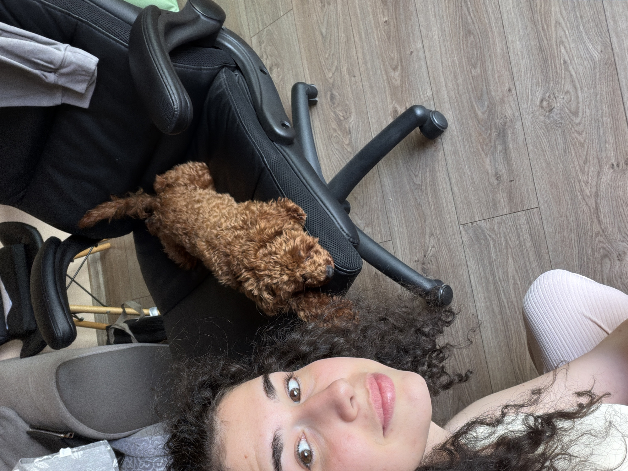 A young woman with dark curly hair and brown eyes is lying down, looking at the camera with a slight smile. She is wearing a light-colored top and pale pink ribbed pants. Next to her, a small, reddish-brown poodle is sleeping on a black office chair. The chair has armrests and a five-wheeled base. In the background, a portion of a gray sofa is visible. The scene is set on a wooden floor. There are no indications of the location being in Sofia, Bulgaria.
