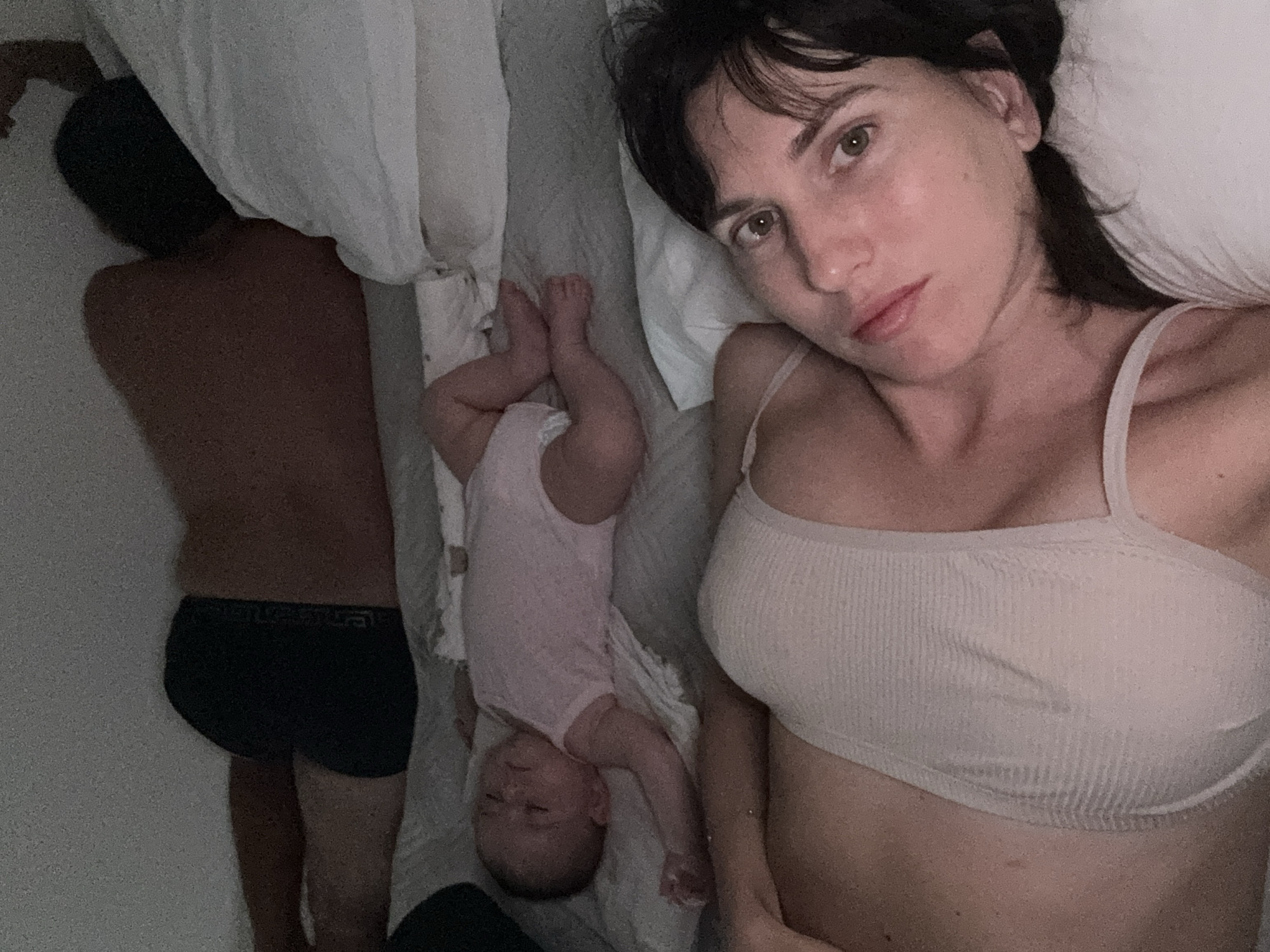 In a dimly lit bedroom, likely in Isola delle Femmine, Italy, three individuals are resting on a bed. In the foreground, a woman with dark hair is lying on her side, facing the camera. She is wearing a light-colored bra. To her left and further back, a baby is lying on its back, dressed in a pink onesie. Beyond the baby, an adult, presumably male given the visible underwear band, is lying on his side with his back to the camera. The bed appears to be made with grey sheets and white pillows. The overall impression is one of a family settling in for rest or sleep.