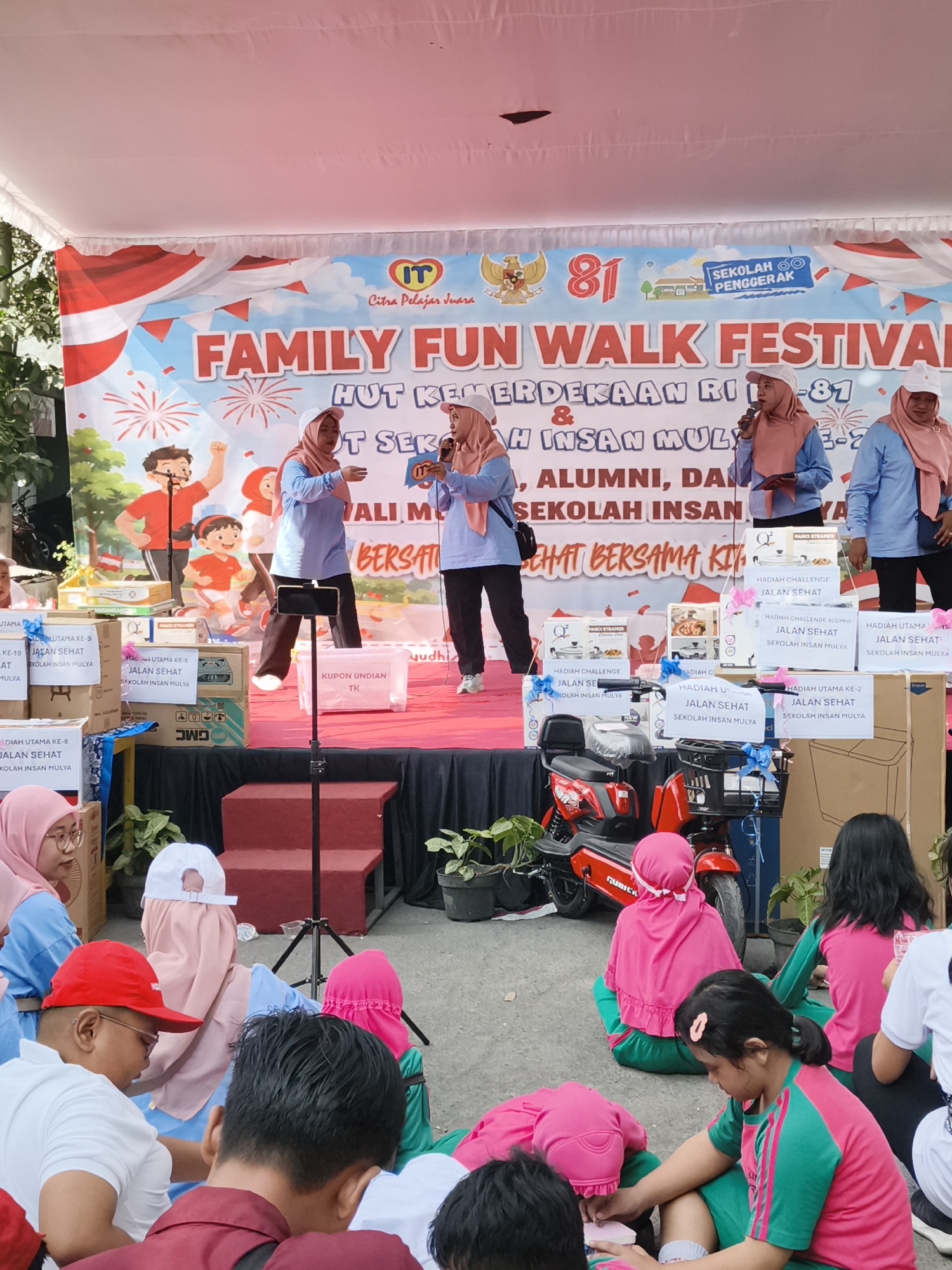 In Petiken, Indonesia, a vibrant community event is unfolding. A banner at the back proclaims "FAMILY FUN WALK FESTIVAL HUT KEMERDEKAAN RI - 81" and "SEKOLAH INSAN MULYA." Three women in headscarves and long-sleeved shirts are on a stage, one speaking into a microphone. Two are wearing light blue shirts and the other a blue and beige patterned shirt.
Below the stage, several people are seated on the ground. A group of children in bright pink and green attire are in the foreground. One girl in the center is drawing on a piece of paper, while another child next to her is using a tablet. A boy wearing a red cap and glasses is in the bottom left, looking down at a piece of paper.
Various boxes are stacked on the stage, some labeled with prize information like "HADIAH UTAMA" (Grand Prize) and "JALAN SEHAT" (Healthy Walk). A red electric motorcycle is parked on the side of the stage. The ground is a paved area, and potted plants are visible around the stage. The overall atmosphere suggests a celebratory gathering with a focus on family and community.