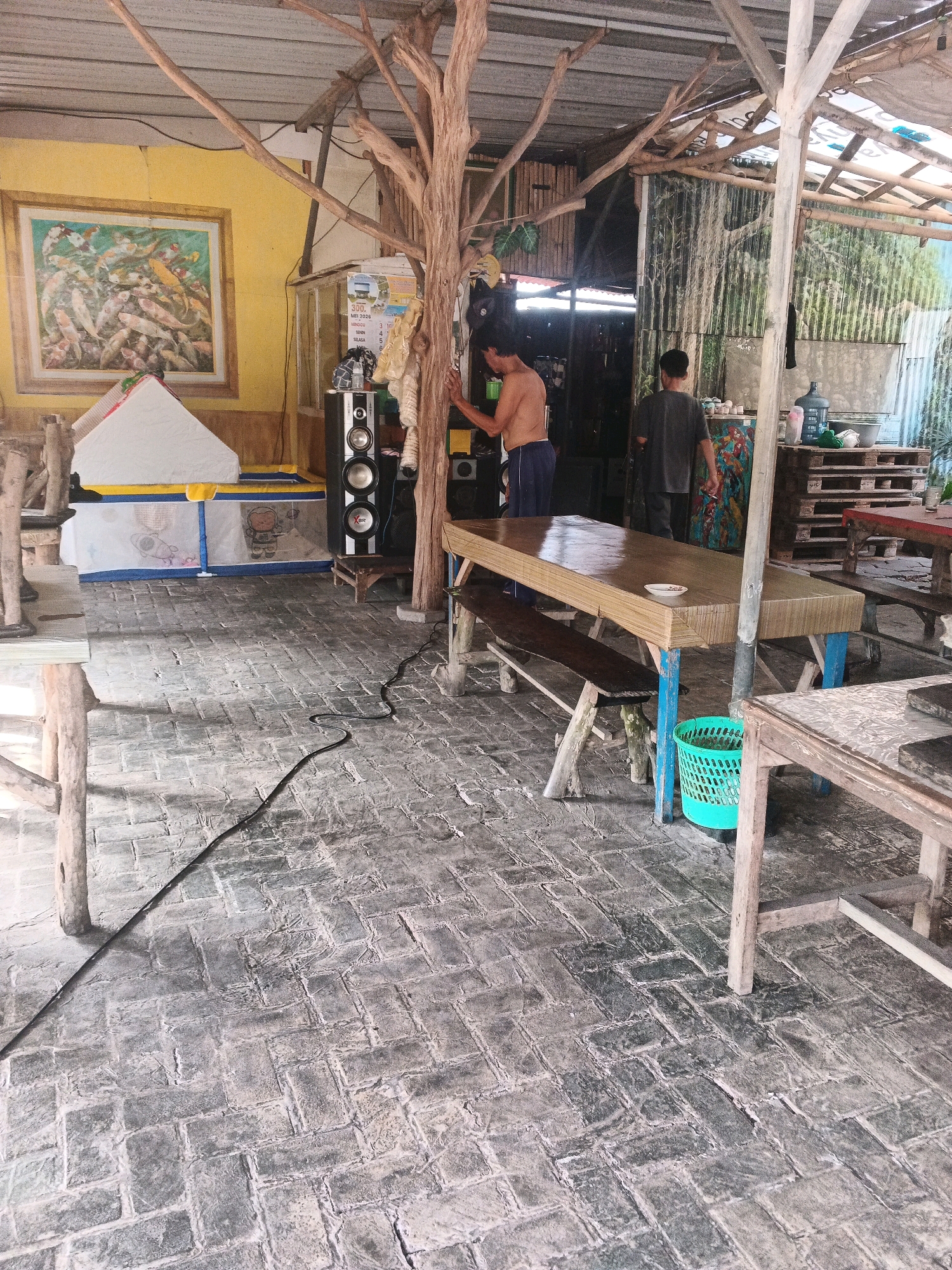 The image depicts an open-air dining area with a rustic aesthetic, likely located in Ungaran, Indonesia. The floor is paved with grey interlocking bricks. In the foreground, a wooden table with a woven surface and blue legs is set up, accompanied by a wooden bench. To the right of this table, another wooden table with a patterned surface is partially visible. A bright green plastic basket sits beneath the woven table.
In the mid-ground, a man without a shirt, wearing dark blue shorts, stands near a large speaker system. He appears to be interacting with or near a wooden structure. Further back, another man wearing a dark grey t-shirt and trousers is walking away from the camera, towards the right side of the image. Behind these individuals, a large painting of koi fish hangs on a yellow wall. Various items, including a speaker, a glass display cabinet with a calendar, and what appears to be a water cooler, are present.
The environment suggests a casual, possibly outdoor eatery or gathering space. The ceiling is constructed of corrugated metal and wooden beams. The overall impression is one of a simple, functional, and somewhat cluttered space.