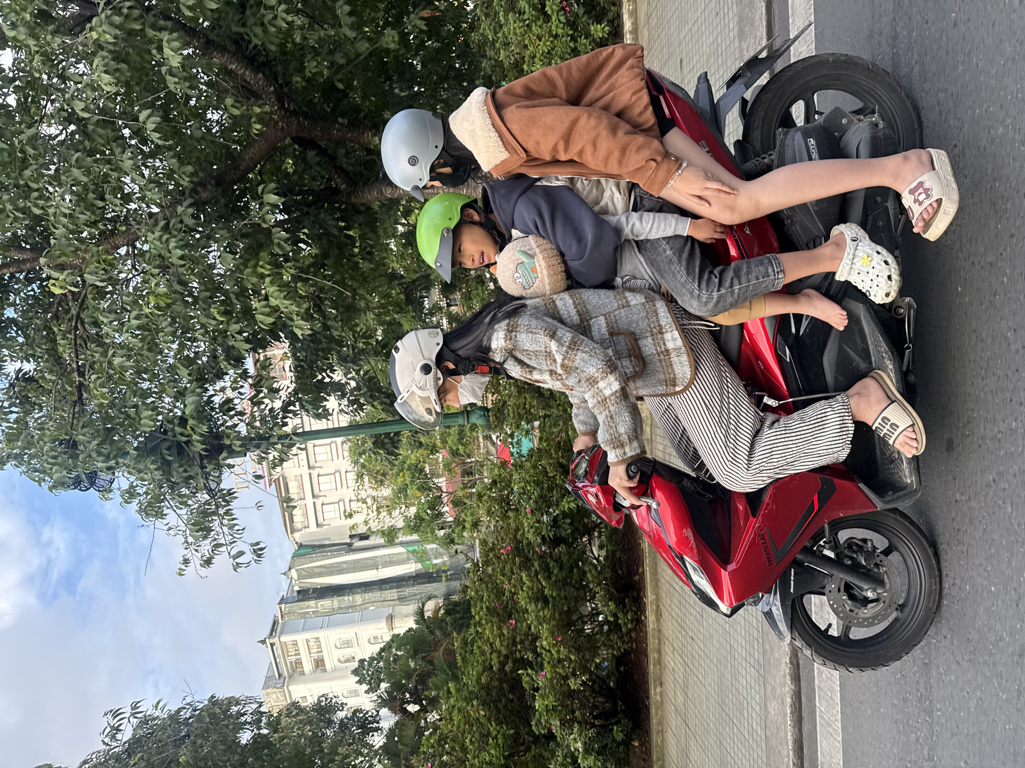 A family of three is riding on a red Honda scooter on a paved street in Da Lat, Vietnam. The mother, wearing a plaid jacket and striped pants, is driving. She has a white helmet and a face mask. A boy, wearing a green helmet and holding a stuffed dinosaur, is sitting in the middle. A girl, wearing a brown jacket and a light blue helmet, is sitting in the back. She is wearing a bracelet. The background shows lush green trees and a multi-story building. The sky is partly cloudy.