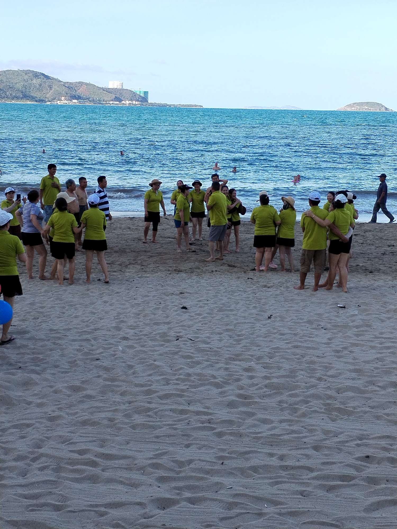 The image shows a group of approximately 25 people gathered on a sandy beach at Bac Nha Trang Ward, Vietnam. They are wearing similar green shirts and a mix of shorts and skirts. The group is in the foreground, seemingly engaged in conversation or an activity, with some individuals standing close to each other. Behind them, the ocean stretches to the horizon, with some indistinct figures swimming or standing in the water. In the background, there's a land mass with buildings and greenery. The sky is light blue. A man in dark clothing is walking along the beach.