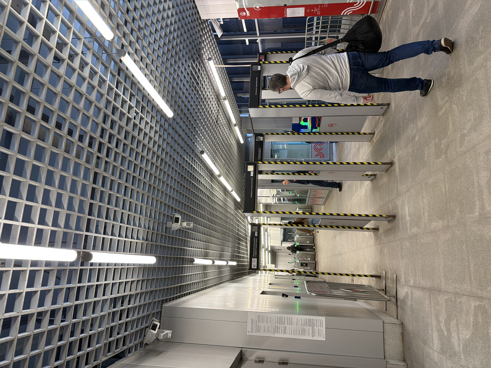 A man in a grey hoodie and blue jeans walks away from the camera and towards a security checkpoint with a black bag slung over his right shoulder. He is passing through an open doorway of a metal security gate with yellow and black striped poles. The security gate is part of a series of similar gates that appear to be a security screening area. Ahead of him, other people are visible passing through similar gates. The environment appears to be an indoor public space, possibly a transportation hub, with a modern, industrial design. The ceiling is a grid structure with fluorescent lights. The floor is tiled with large grey tiles. There are security cameras mounted on the ceiling. Signs are visible on the walls, some with text in a foreign language.