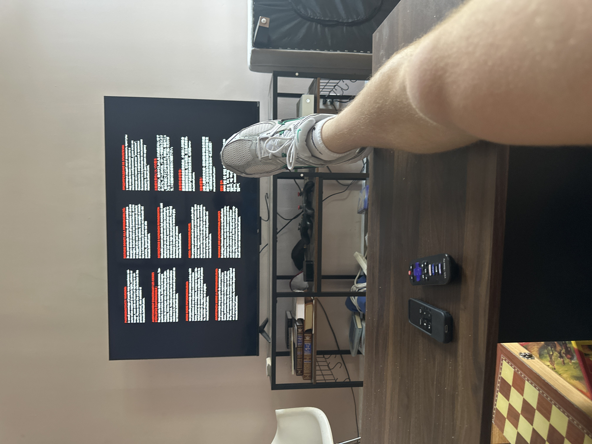 A horizontal, eye-level indoor shot captures a casual scene featuring a person's lower leg and foot, a television screen displaying text, and a desk with household items. The image itself is rotated 90 degrees counter-clockwise.
In the foreground, a Caucasian person's bare lower leg and foot are visible. The foot is clad in a white athletic sneaker with green accents and white ankle socks, and is positioned above a dark wooden desk or table. The sneaker has a mesh upper and white laces.
On the desk, two black remote controls are present; one is identifiable as a Roku remote due to its branding and purple button. A decorative wooden box with a yellow and red checkered pattern and an equestrian image on its side is partially visible on the desk.
The central subject is a large, dark television or monitor screen on the left, displaying multiple blocks of white text on a black background, with certain words highlighted in red. The text comprises warning messages in various languages, likely related to photosensitivity. Visible phrases include "PHOTOSENSITIVITY WARNING" in English, along with warnings in Japanese, Russian ("ОСТОРОЖНО: О СУДОРОГАХ"), French ("AVERTISSEMENT CONCERNANT LES CRISES"), German ("WARNUNG VOR LICHTEMPFINDLICHKEIT"), and Spanish ("ADVERTENCIA SOBRE LA FOTOSENSIBILIDAD"). The red text typically introduces each warning block or emphasizes key terms.
Behind the screen and desk, a dark metal shelving unit holds several items, including ornate, dark-bound books, and various cables and electronic accessories. Above the shelving unit, a dark grey upholstered object, possibly a futon or bed frame, is visible. The room features light-colored walls. The setting suggests a casual, residential environment, possibly a living room or bedroom. The activity appears to be relaxing, possibly watching media or playing a video game, given the nature of the warning screen. No specific time of day or weather conditions are discernible from the indoor scene.