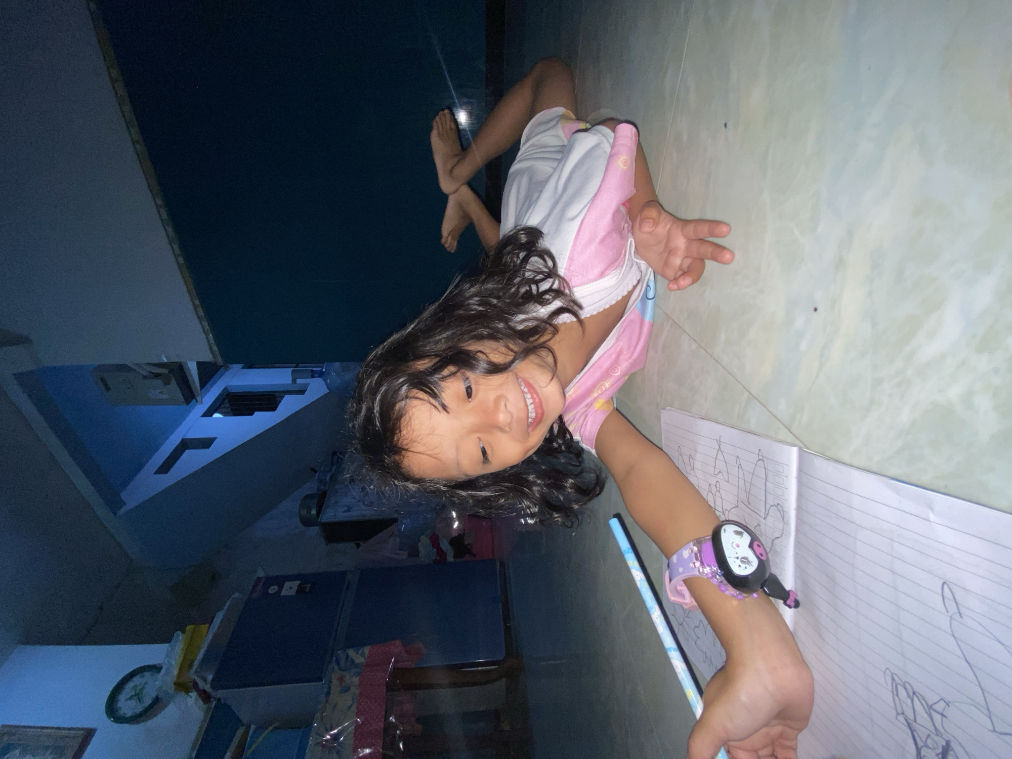 In the city of Gelam, Indonesia, a young girl with dark hair lies on her stomach on a tiled floor, engaged in drawing in a notebook. She is wearing a pink and white garment. Her right arm is extended, holding a light blue pencil with a cartoon character design. A purple watch with a black face and a pig-like character is visible on her wrist. The notebook contains sketched drawings. The background shows a dimly lit interior with blue walls, a staircase, and various objects, including a clock on a shelf and what appears to be a refrigerator covered with plastic. The overall lighting suggests it is nighttime or the room is dimly illuminated.