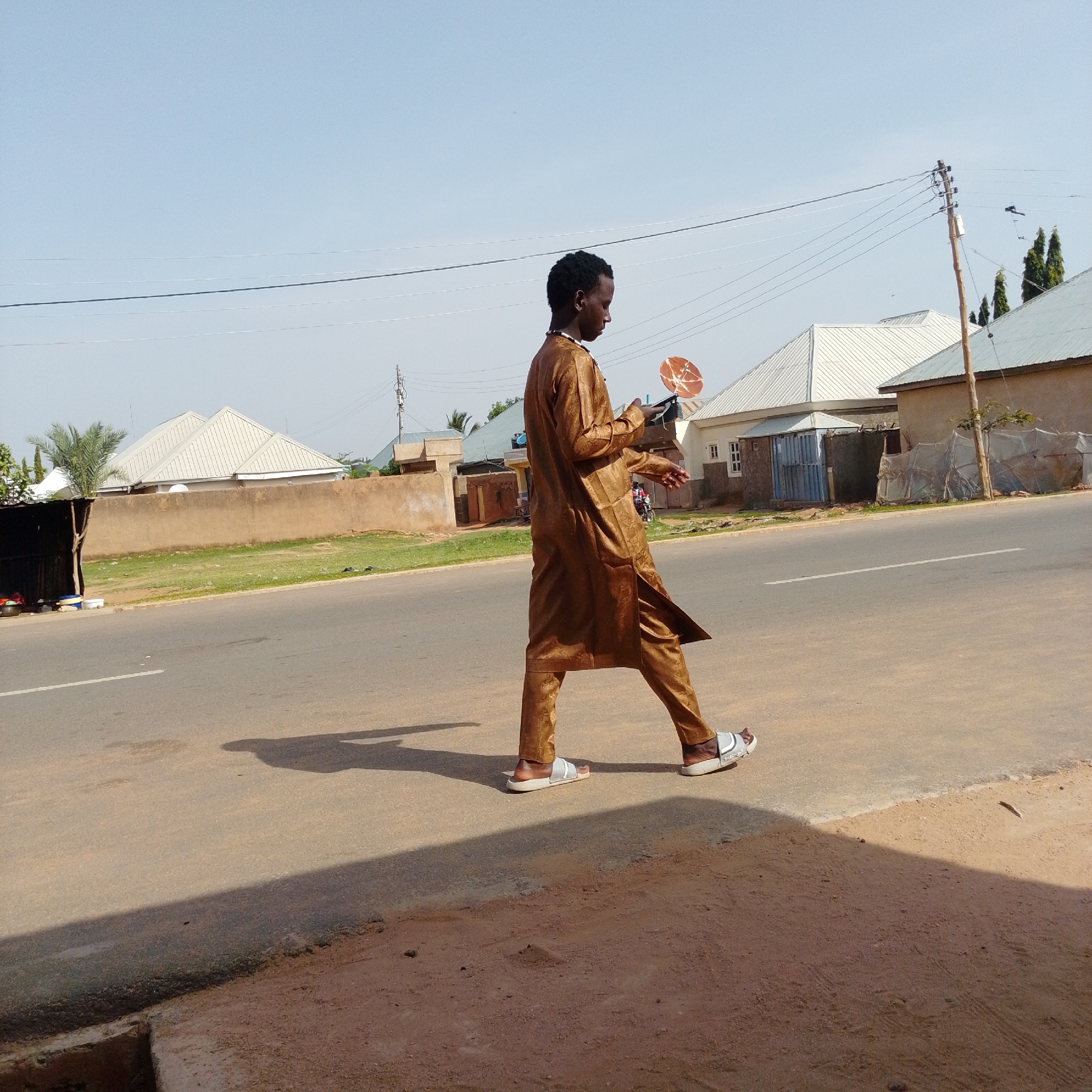 A young man in a golden brown robe and white sandals walks along a paved road, looking at a dark electronic device in his hands. He is in profile, walking from left to right. His shadow is cast long on the road beside him. In the background, there are houses with metal roofs, a beige concrete wall, green grass, and trees, suggesting a residential area. Power lines and a utility pole are visible against a clear blue sky. The scene appears to be in a city or town in a warm climate.