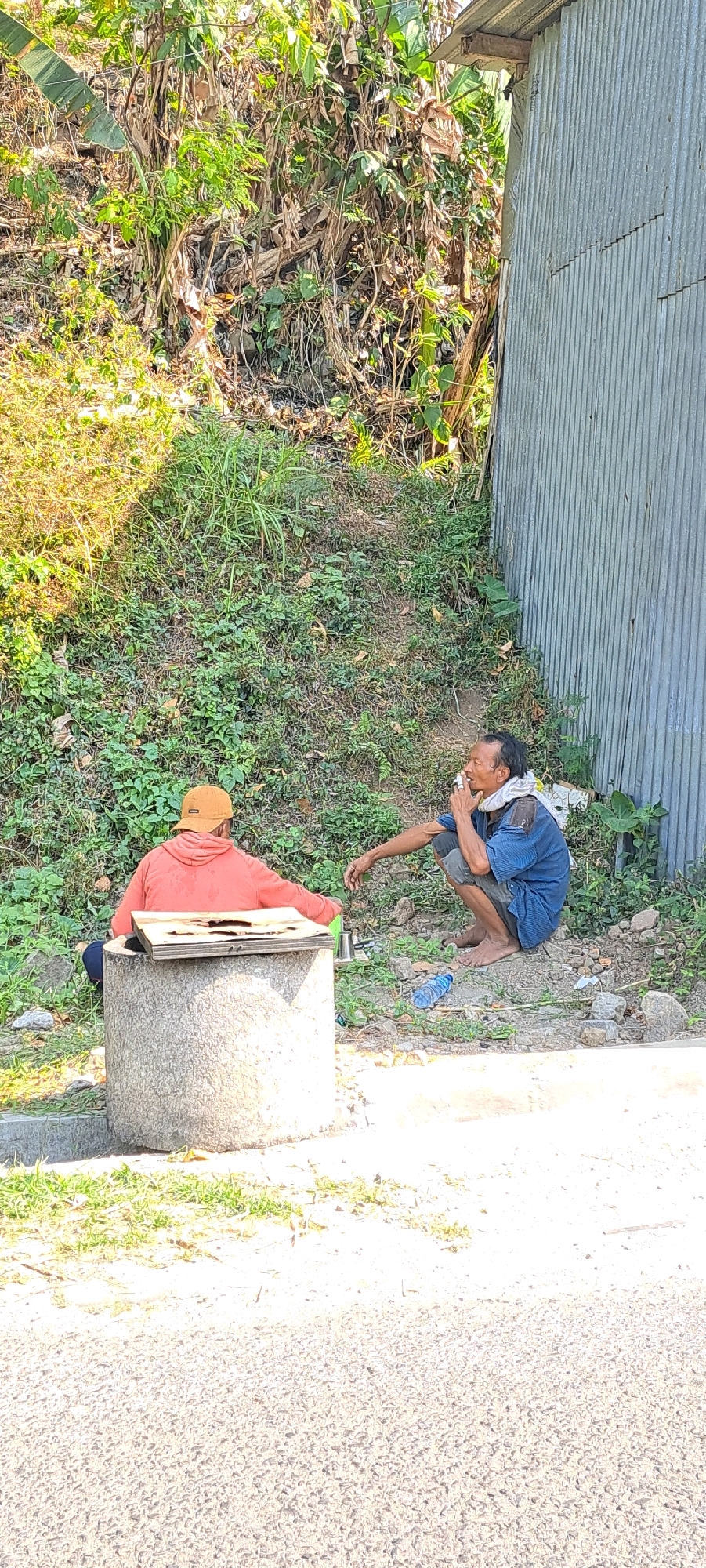 In Watuagung, Indonesia, two individuals are situated near a corrugated metal structure and a concrete well. One person, facing away from the camera, is seated on the well, wearing an orange hooded jacket and a cap, with their hands resting on a wooden plank placed on the well's rim. The second individual is crouched down to the right, facing left and smoking a cigarette. This person wears a blue shirt, grey shorts, and a white cloth around their neck. A plastic water bottle lies on the ground between them. The environment is characterized by lush green vegetation and the presence of a dirt and gravel pathway in the foreground.