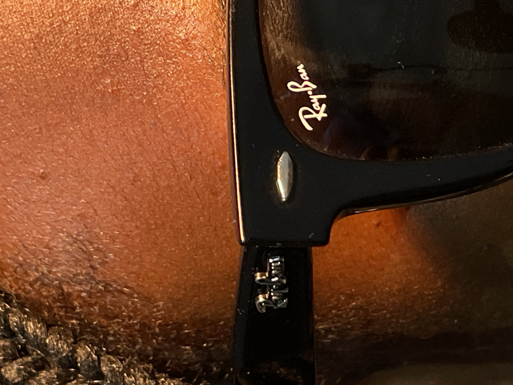 The image shows a close-up of the right side of a pair of black Ray-Ban sunglasses resting on a person's skin. The Ray-Ban logo is visible on the lens. The temple of the sunglasses is also visible, along with a silver accent on the frame. The person's skin tone is warm, and there is hair visible near the bottom of the frame.
There is no information that this image was taken in the city of Barakin Akawo, Nigeria.