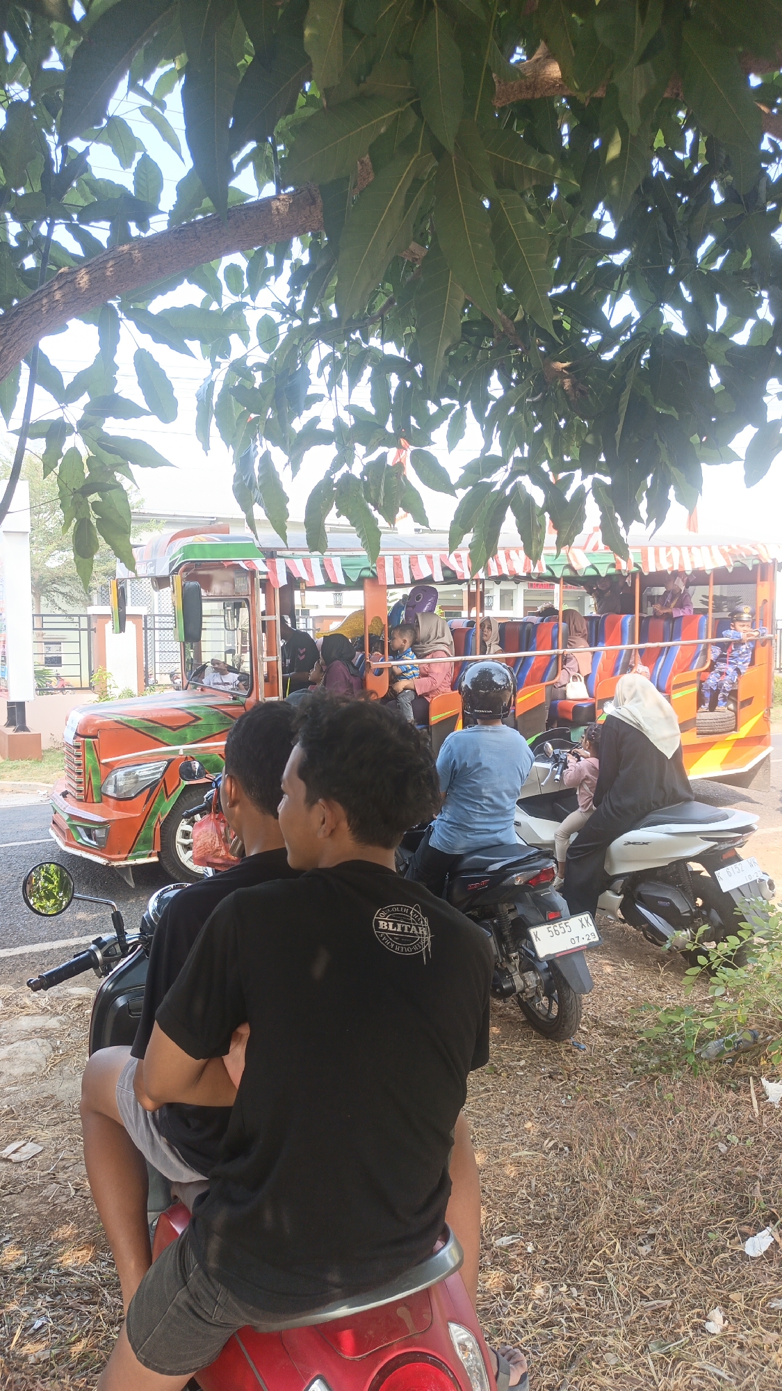 In the city of Kandangmas, Indonesia, two young men are seated on a red scooter. The passenger in the back wears a black t-shirt with white lettering. The driver, facing away, is wearing a light blue t-shirt and a black helmet. They are positioned in the foreground, with a brightly colored bus-like vehicle behind them. The bus, decorated with orange, green, and red stripes, carries several passengers, including women wearing headscarves and a child. The vehicle appears to be a form of public transportation. The scene is set outdoors, with a dirt and dry grass ground cover. Lush green tree leaves and branches overhang the scene from above, suggesting it is daytime and possibly a sunny day due to the dappled light. In the background, a building with white walls and a fence can be seen. The overall impression is a snapshot of everyday life and transportation in a tropical environment.