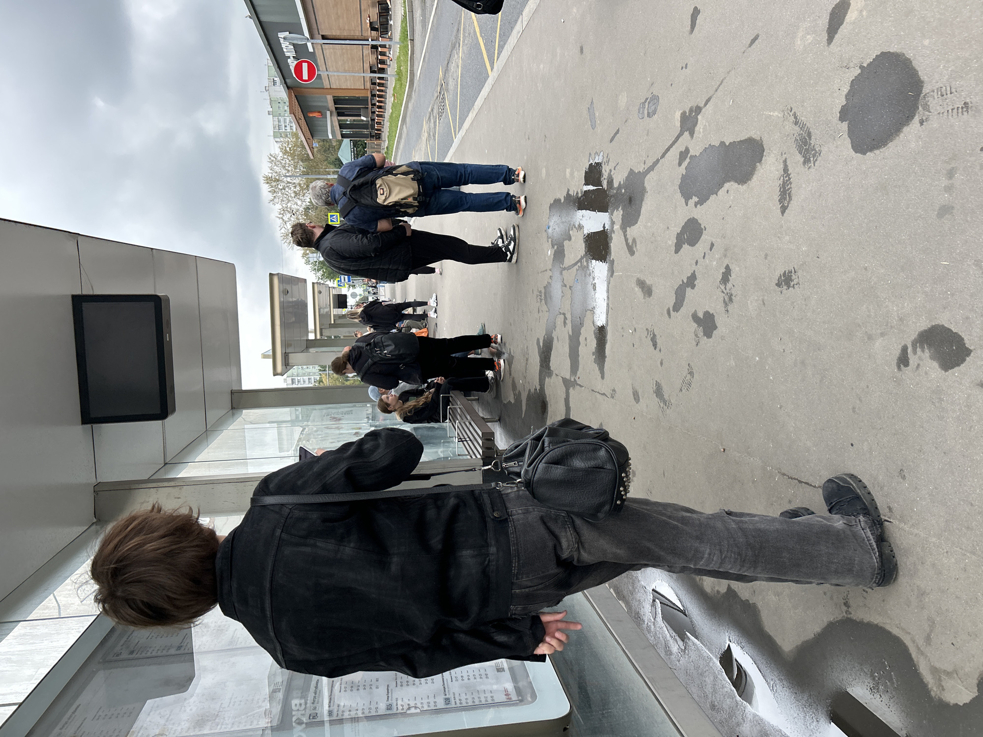 The image depicts a scene at a bus stop in Moscow, Russia, under an overcast sky. In the foreground, a person with brown, shaggy hair, wearing a black denim jacket and dark grey jeans, stands with their back to the camera, holding a phone. A black leather handbag with studded accents hangs from their shoulder. The bus stop features a glass shelter with a timetable displayed on a panel. To the right, a bench is occupied by several people, including a young woman and a child. Further down the bus stop, two men are conversing. One man, wearing a black jacket and trousers, has his arm around the other man, who is wearing a blue shirt, jeans, and a beige backpack. In the background, a McDonald's restaurant is visible with a red prohibition sign. The ground is damp with puddles and visible footprints, suggesting recent rain. The environment appears to be an urban street with buildings and trees lining the background.