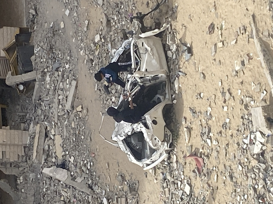 In Khan Yunis, Palestinian Territory, a wide shot captures a scene of urban destruction under a dull sky. Two individuals are situated around a severely damaged white sedan, which occupies the foreground-middle ground. The car is extensively crumpled, with its roof collapsed inwards, windows shattered, and body panels warped and torn.
An adult, dressed in dark clothing that includes a head covering, is seated partially inside the vehicle on the left side, appearing to reach towards the car's interior. A younger individual, possibly a child or adolescent, wearing a blue and white hooded jacket and dark pants, is perched on the collapsed roof or rear trunk area of the car, facing inwards and also interacting with the vehicle's remains. Both individuals are focused on the wrecked vehicle, their exact actions undetermined but suggesting examination or salvage.
The ground surrounding the car is a barren landscape of tan sand heavily interspersed with a dense field of concrete rubble, fragmented building materials, and debris. In the background, further piles of structural debris and remnants of destroyed buildings are visible, indicating widespread damage to the urban environment.