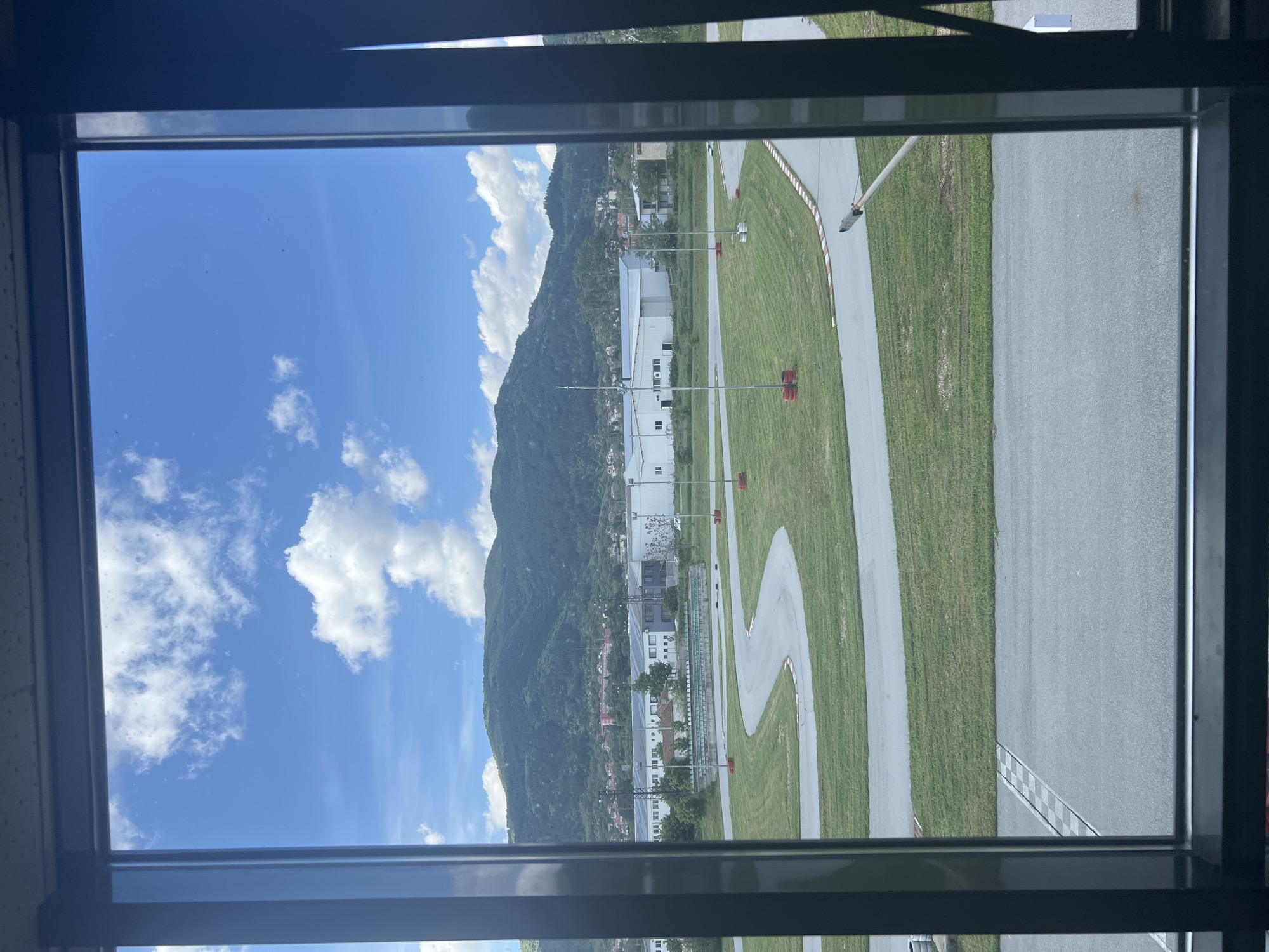 View from a window of a landscape including a mountain and a racetrack in Tvarditsa, Bulgaria. The sky is blue with scattered cumulus clouds. The mountain is densely forested. Buildings are located at the base of the mountain. A grey racetrack with painted white lines and red and white barriers winds through a green grassy field. The window frame is dark grey and obscures parts of the view.