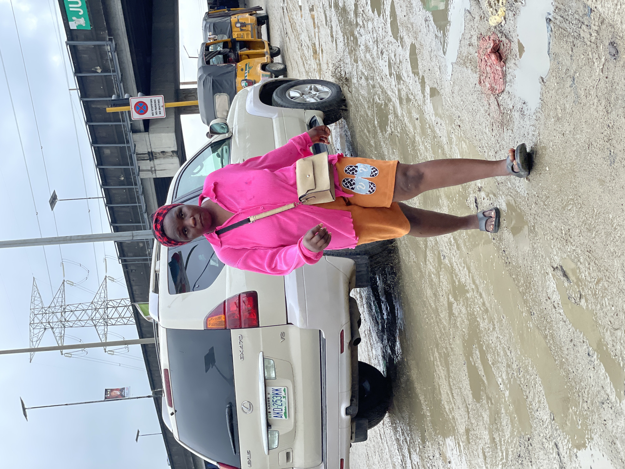 In Lekki, Nigeria, a woman walks on a muddy, waterlogged road, her dark skin partially obscured by a vibrant pink, long-sleeved, collared shirt and orange shorts. The shorts are adorned with a white slipper print. A beige crossbody bag rests on her left hip. She wears dark flip-flops. Behind her, a beige Lexus GX 470 SUV is parked, its license plate reading "AKD-253KK". Further in the background, three yellow auto-rickshaws, common in Nigeria, are lined up. Overhead, a large bridge supports a "JUNCTION" sign and a "NO STOPPING NO PARKING NO WAITING" sign. A towering electricity pylon stands prominently on the left, with a flag and a banner on a pole nearby. The sky is overcast.