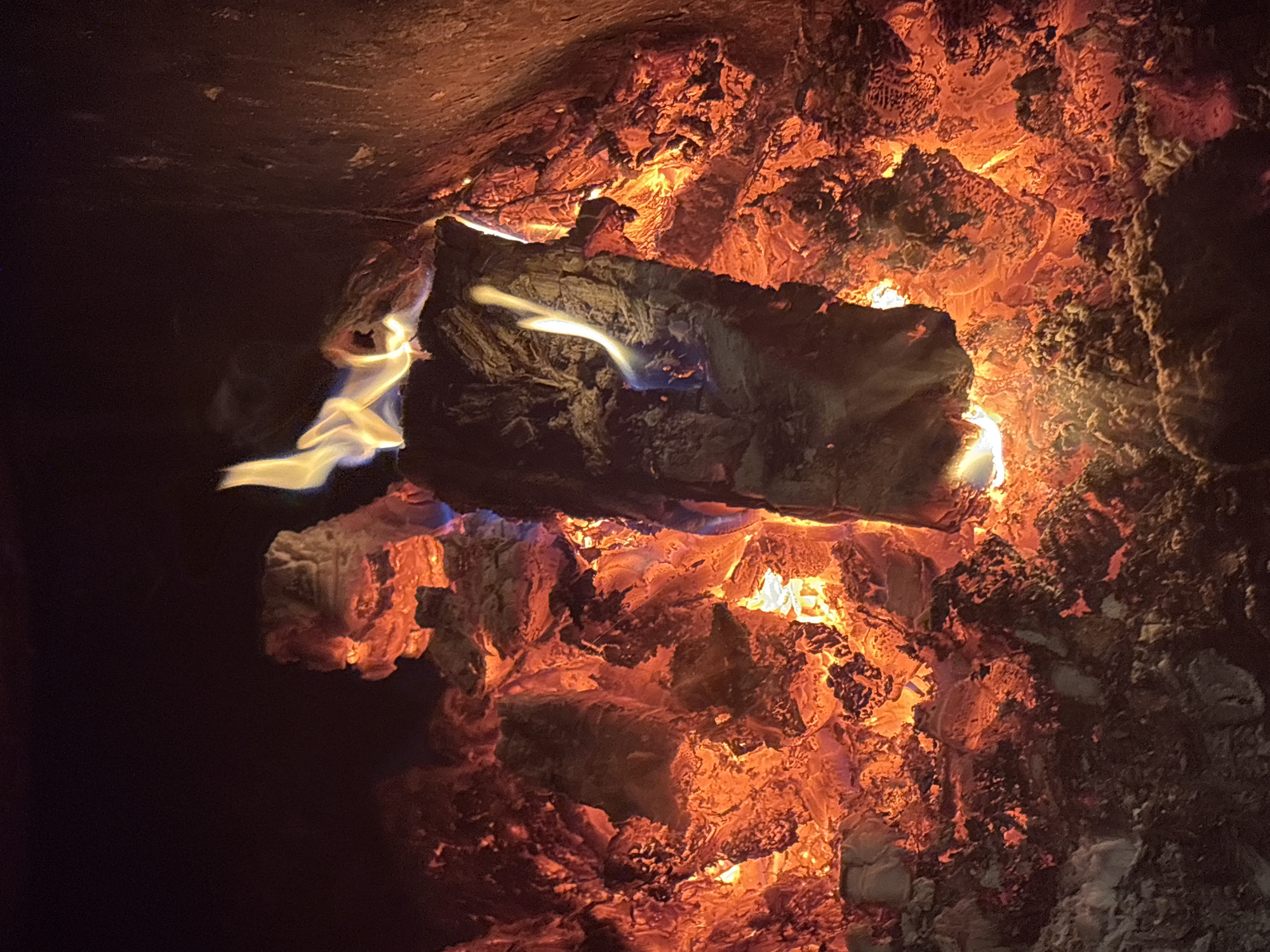 The image depicts a close-up view of a roaring fire. The primary subjects are burning logs and embers, radiating intense orange and red light. Wisps of yellow and blue flames dance above the wood, indicating high temperatures. The background is dark, suggesting the fire is contained within a fireplace or fire pit. There are no people or animals visible in the image. The scene conveys a sense of warmth and intense heat. The location in the city of Курган, Russia is not discernible from the visual information provided.