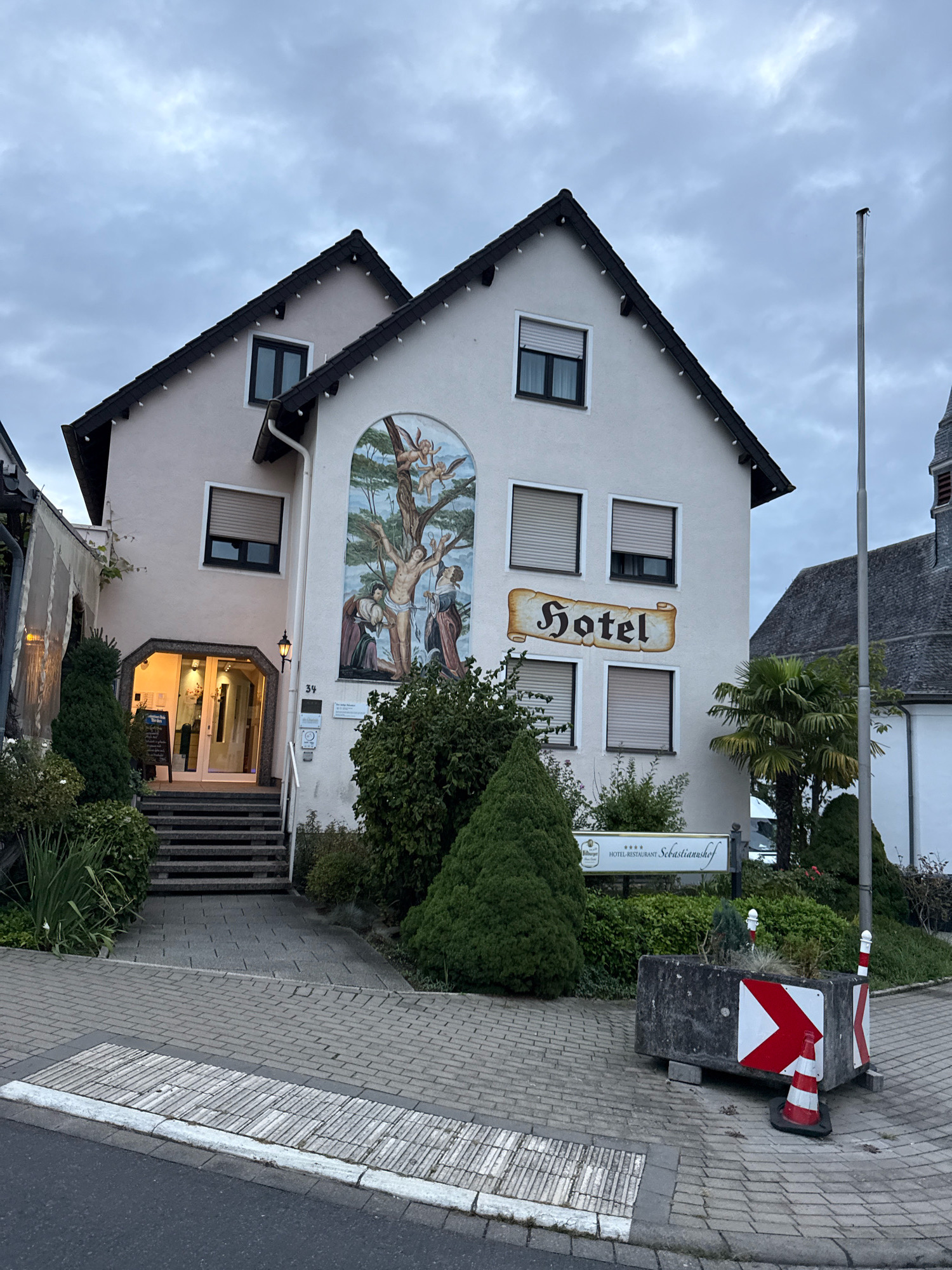 The image shows the facade of a hotel and restaurant named "Sebastianushof" located at Sebastianusstraße 34 in Bonn, Germany. The hotel building is light-colored with a dark roof and features several windows with closed blinds. A large mural depicting a religious scene is painted on the right side of the building. Above the entrance, a sign reads "Hotel" in a stylized font. The entrance area is illuminated and accessible via a set of stone stairs. Surrounding the building are various green plants and trees. In the foreground, a paved walkway leads to the hotel entrance, and a concrete barrier with red and white traffic markings is visible on the right. The sky is overcast, indicating possibly evening or a cloudy day.
