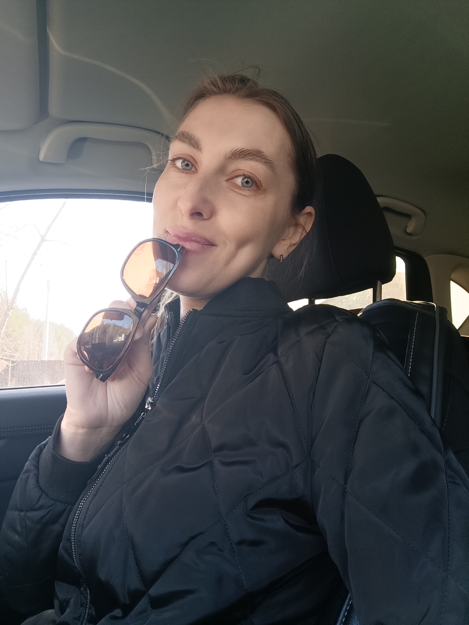 A portrait-style photograph captures a woman seated inside a car, looking directly at the camera with a slight smile. She has light skin, light-colored eyes, and her brown hair is pulled back. She is wearing a black quilted bomber jacket with a zippered front and ribbed cuffs. Her right hand holds a pair of brown-framed sunglasses with orange-tinted lenses, which are playfully positioned near her mouth, resting on her lower lip.
The setting is the interior of a vehicle, identifiable by the grey headliner, a grab handle, and a dark car seat behind her. Through the car window on the left, a bright outdoor scene is visible. Bare trees, suggesting late autumn, winter, or early spring, are prominent, along with what appears to be a light-colored fence or structure in the distance. The natural light indicates it is daytime. The overall mood is casual and relaxed. There is no visible text in the image. The location is contextualized as Chaikovsky, Russia.