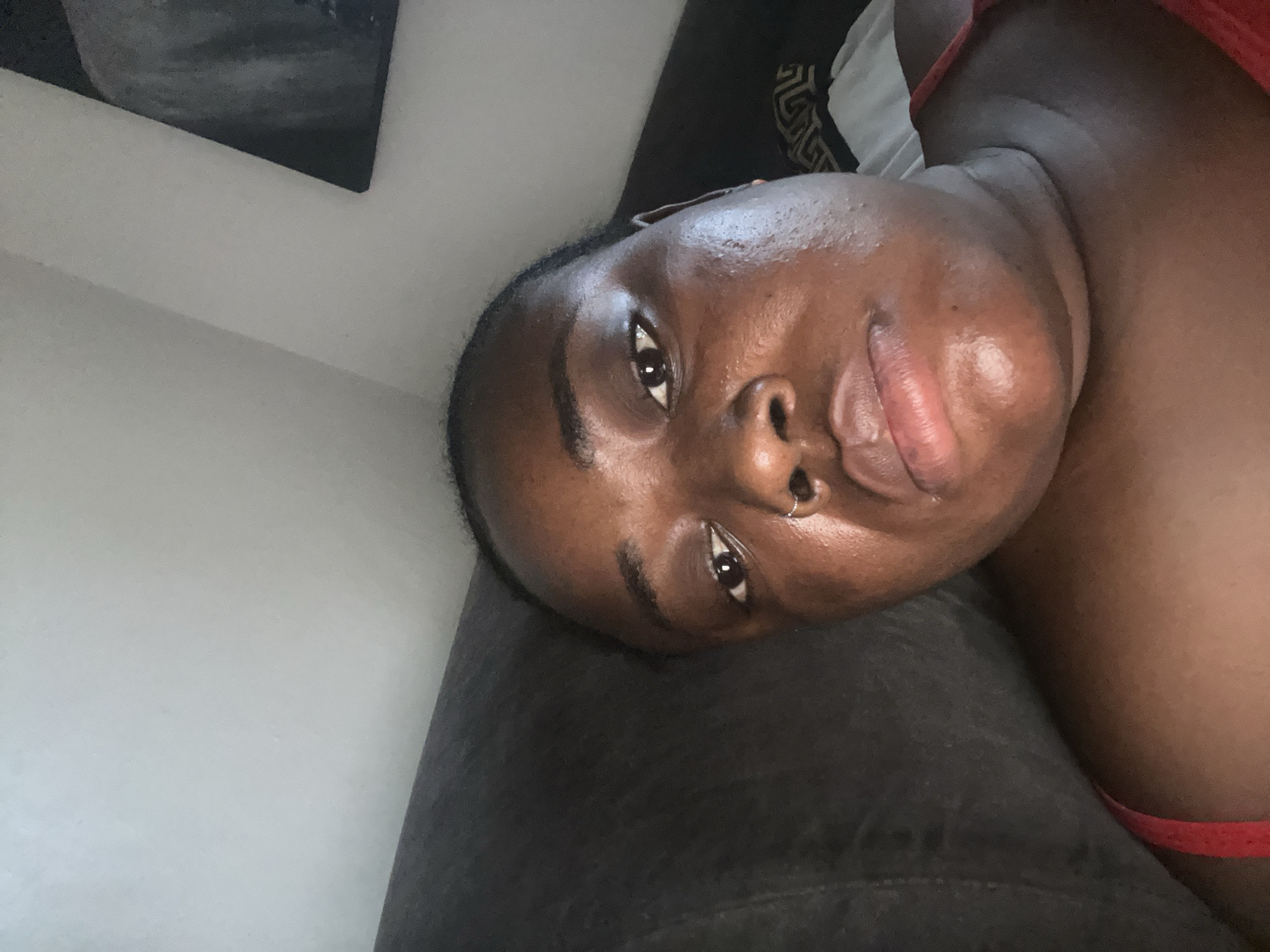 An upward-facing, angled shot captures the face and upper torso of a young adult with dark skin, likely female, resting indoors in Abuja, Nigeria. The individual is positioned on a dark, textured fabric surface, possibly a sofa or bed. Their head is tilted to the right, and their gaze is directed towards the camera with a neutral, relaxed expression.
The person has dark, smooth skin, full lips, and a small silver ring piercing the left side of their nose. Their hair appears dark and styled away from the face. A strap of a red garment, possibly a top or bra, is visible on their right shoulder and chest area. The skin on their face and neck has a slight sheen, suggesting either natural oils or a dewy complexion.
In the background, to the upper left, a light-colored wall is visible, along with the corner of a dark-colored object. This object features a faint, repeating pattern, which appears decorative rather than legible text, possibly resembling a stylized design or motif. The lighting is ambient, indicating an indoor setting. The overall impression is a casual, relaxed pose.